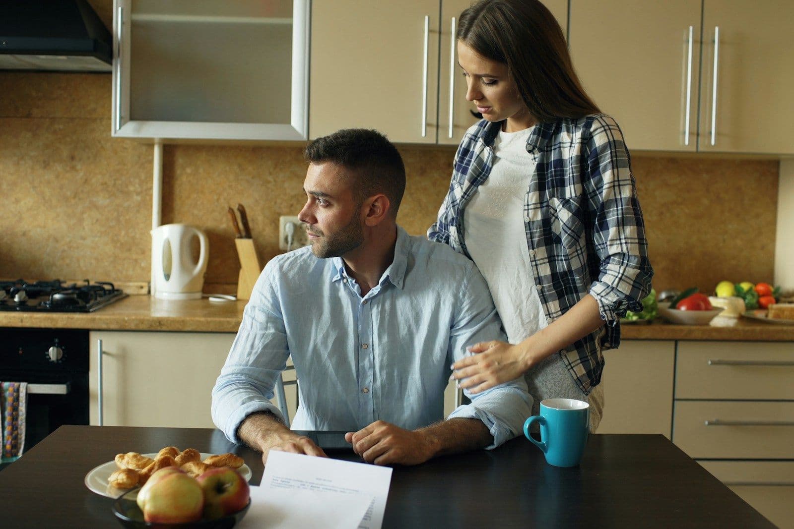 A man sits at a kitchen table with papers while a woman stands beside him, resting a hand on his shoulder.