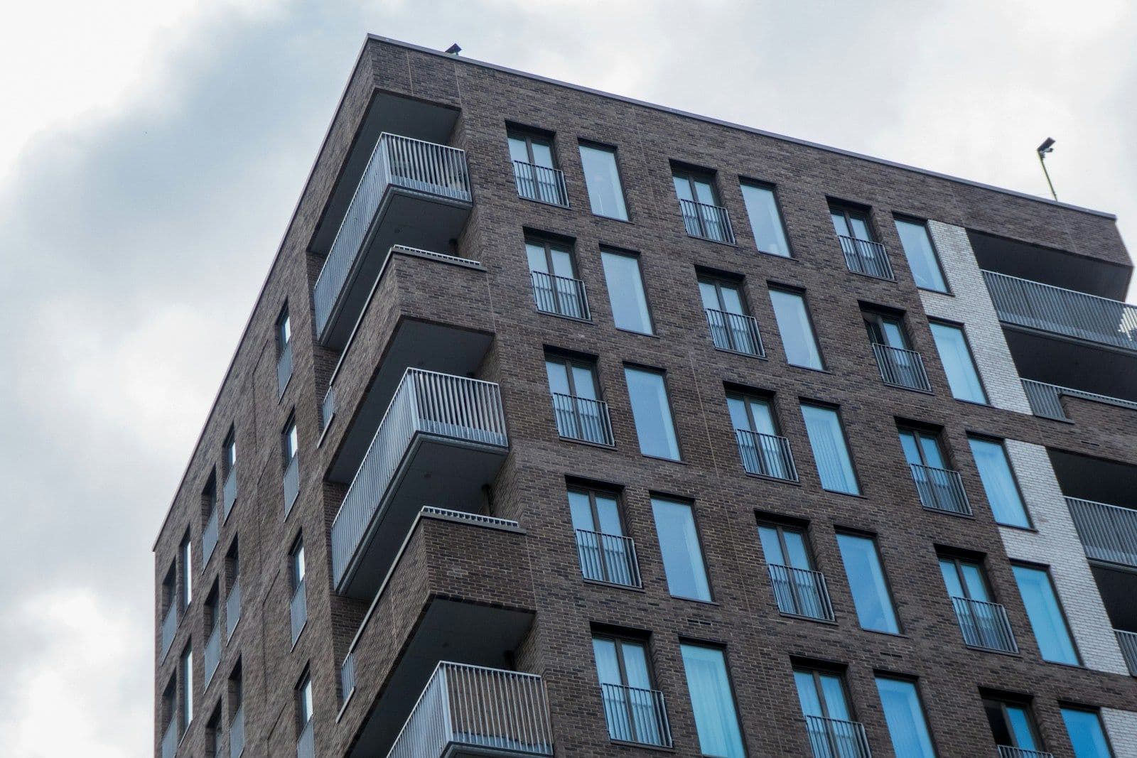 Low-angle view of a multi-story brick apartment building with rows of windows and small balconies under a cloudy sky