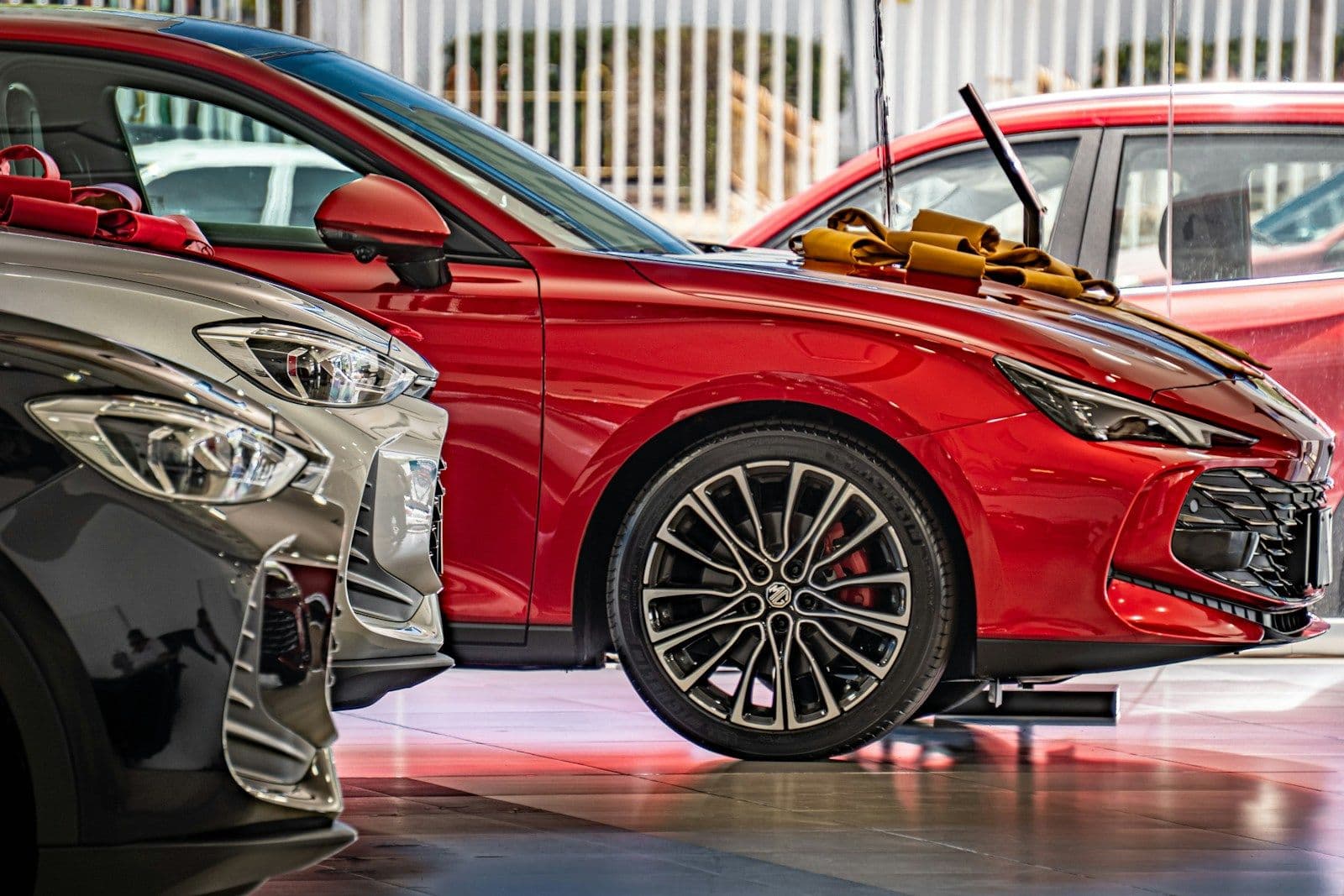 Red sports sedan with silver alloy wheels and red brake calipers displayed in a showroom beside a dark car, with a yellow-tipped car part visible in the background