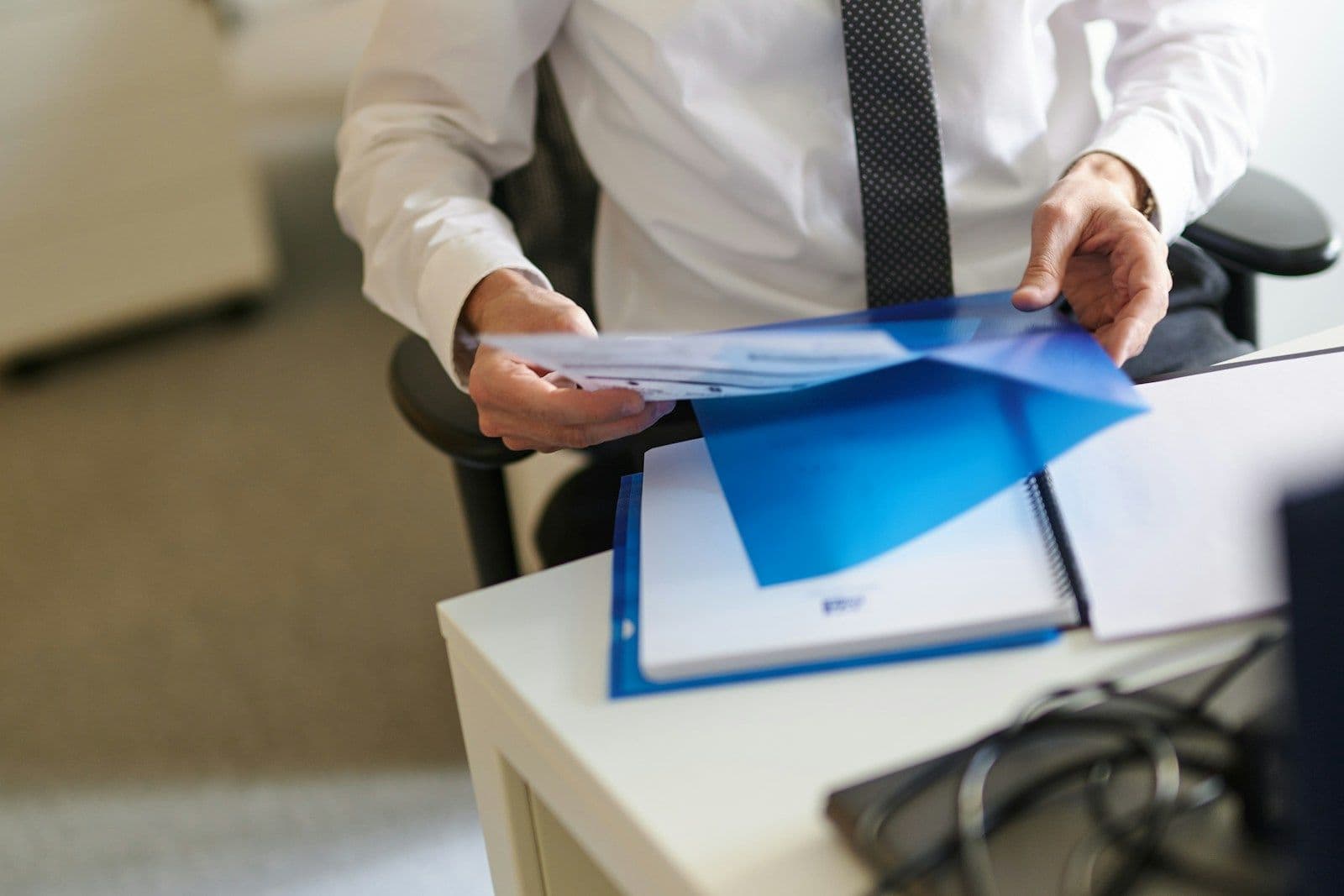 Man in white shirt and dark tie handing a blue folder of papers across a white desk in an office