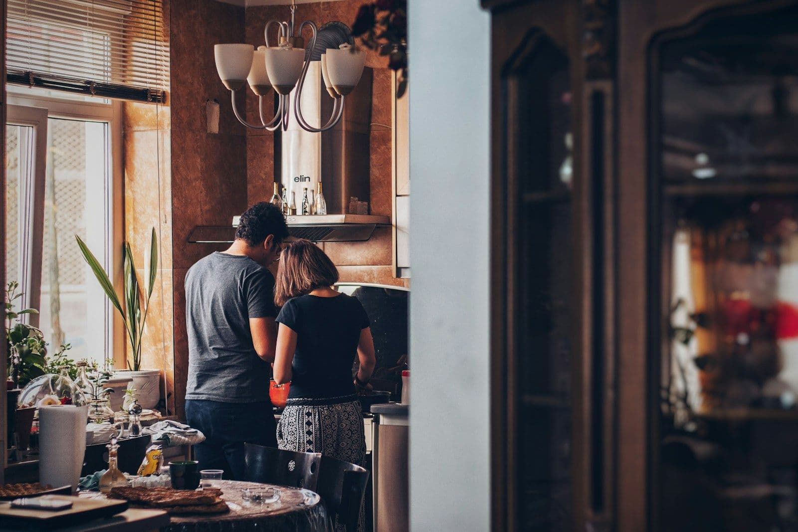 A couple stands together in a warm, softly lit kitchen, seen from behind, in a quiet moment at home.