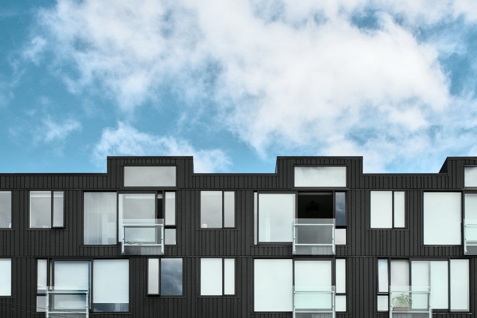 Modern dark-clad apartment building with rows of windows and balconies under a partly cloudy blue sky