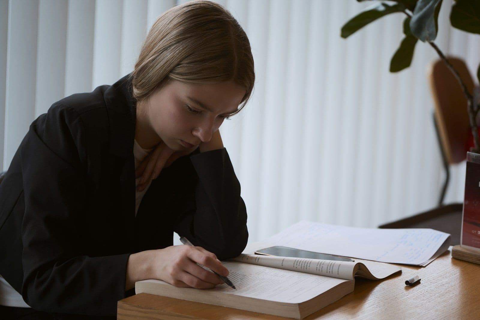 A young woman in a dark blazer writes with a pen in an open notebook at a wooden desk, with papers and a plant nearby.