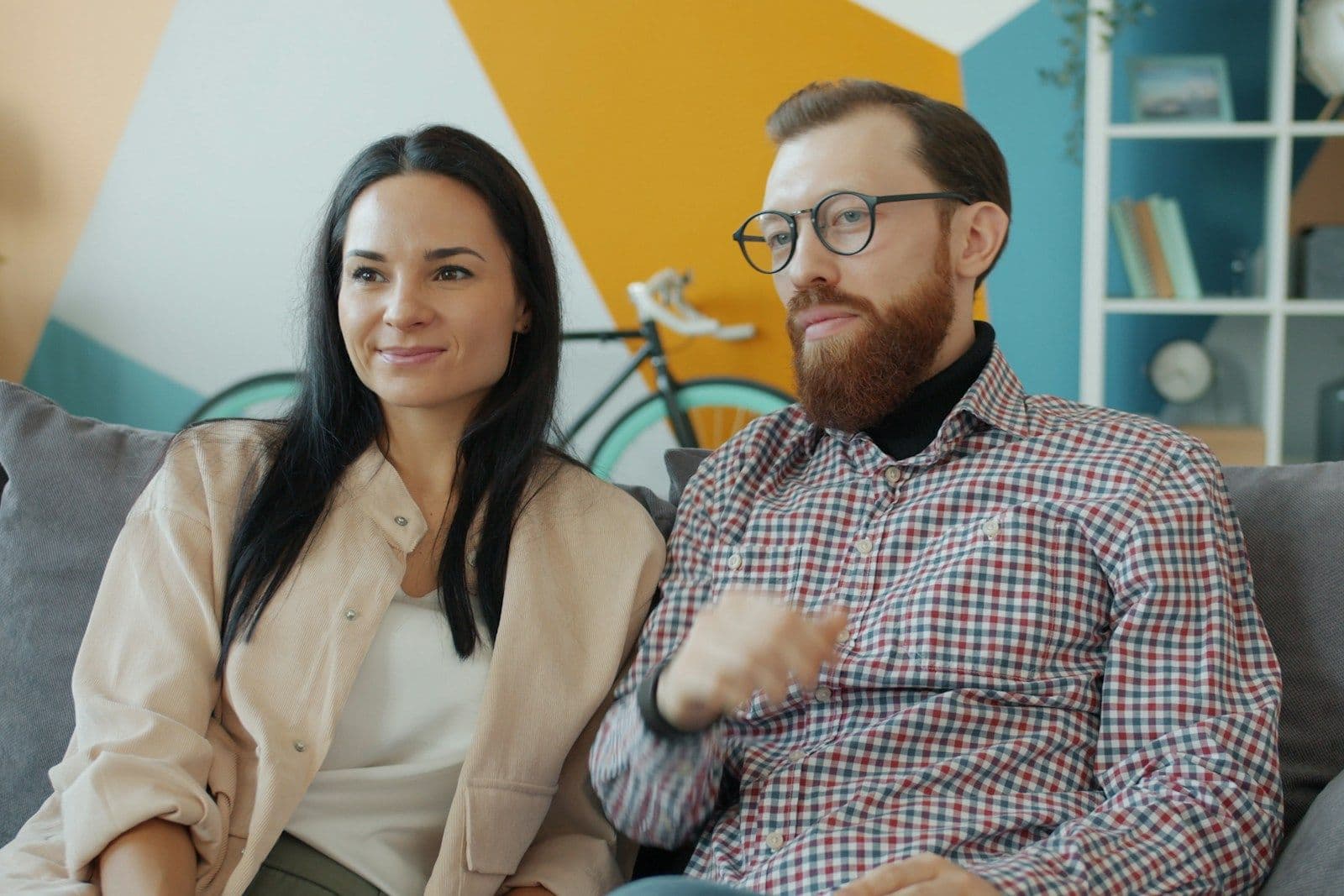 A woman and a bearded man in glasses sit together on a gray sofa in a bright living room, looking ahead calmly.