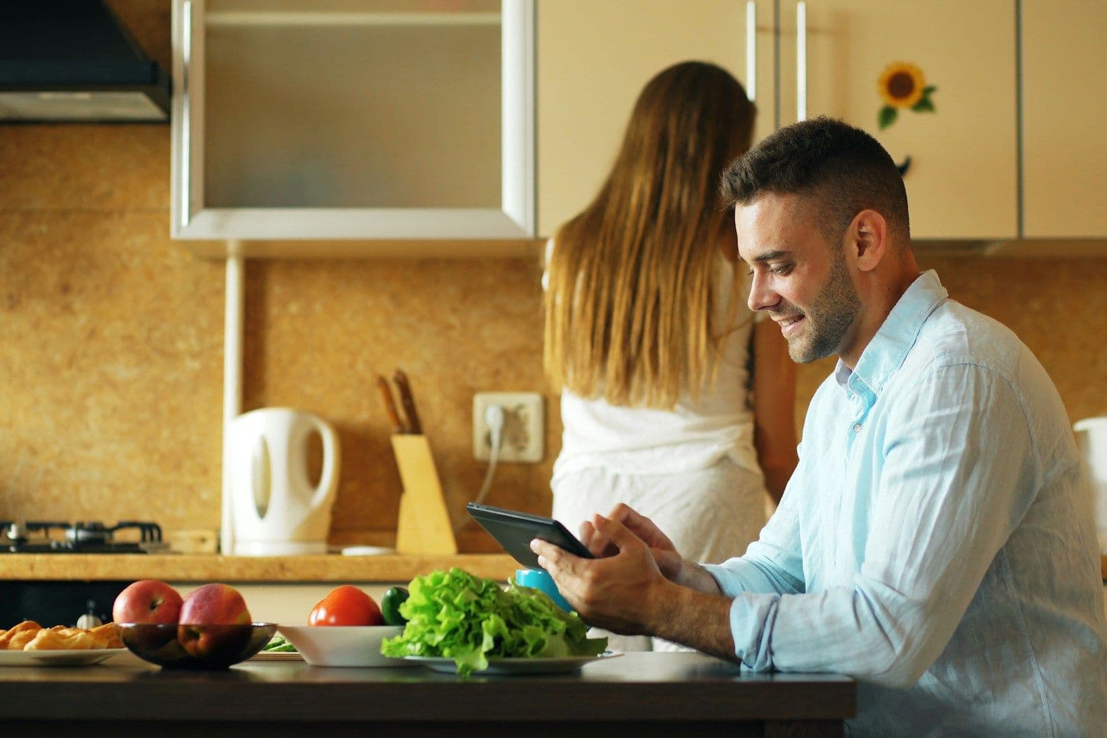 A man smiles while reading a tablet at a kitchen table as a woman stands in the background of a bright home kitchen.