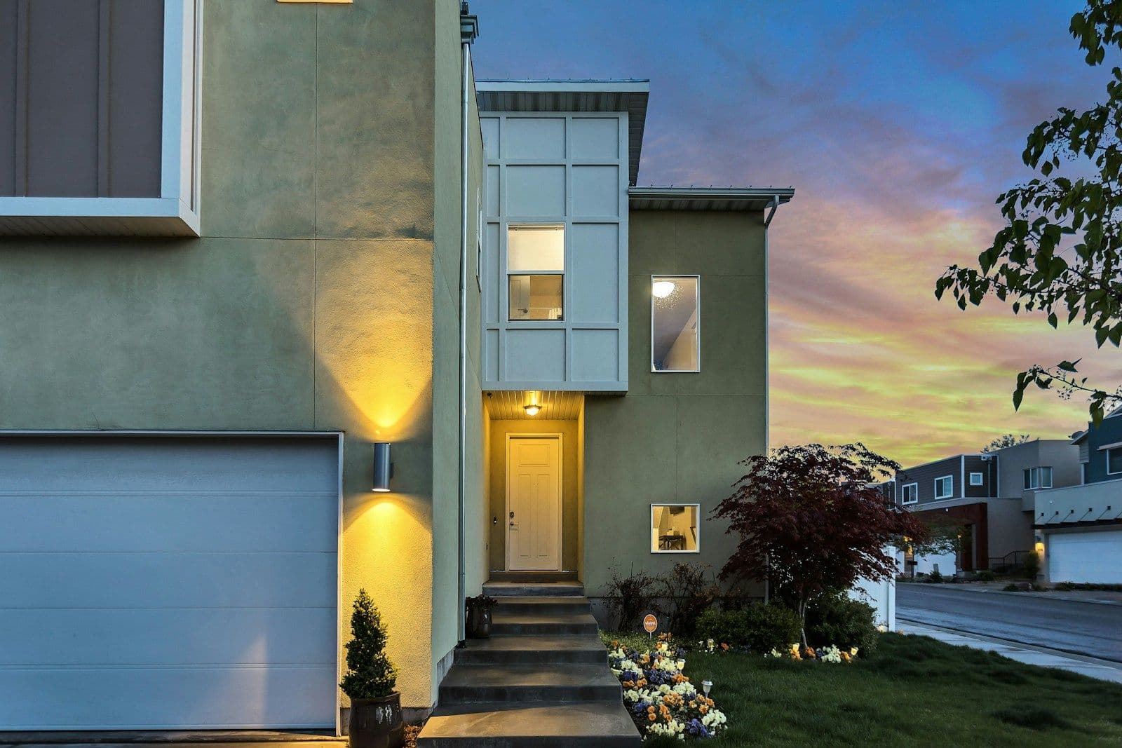 Modern two-story stucco home at dusk with warm wall lights, a white garage door, a lit front entrance, and a pink-and-blue sky.