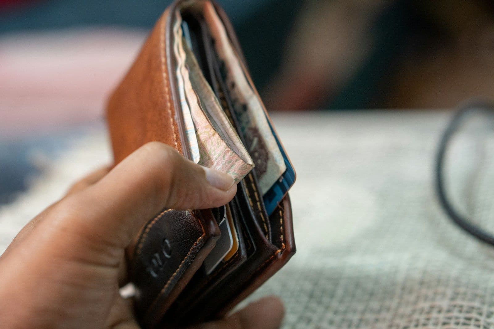A hand holding open a brown leather bifold wallet filled with cards and folded cash, shown in close-up against a blurred background