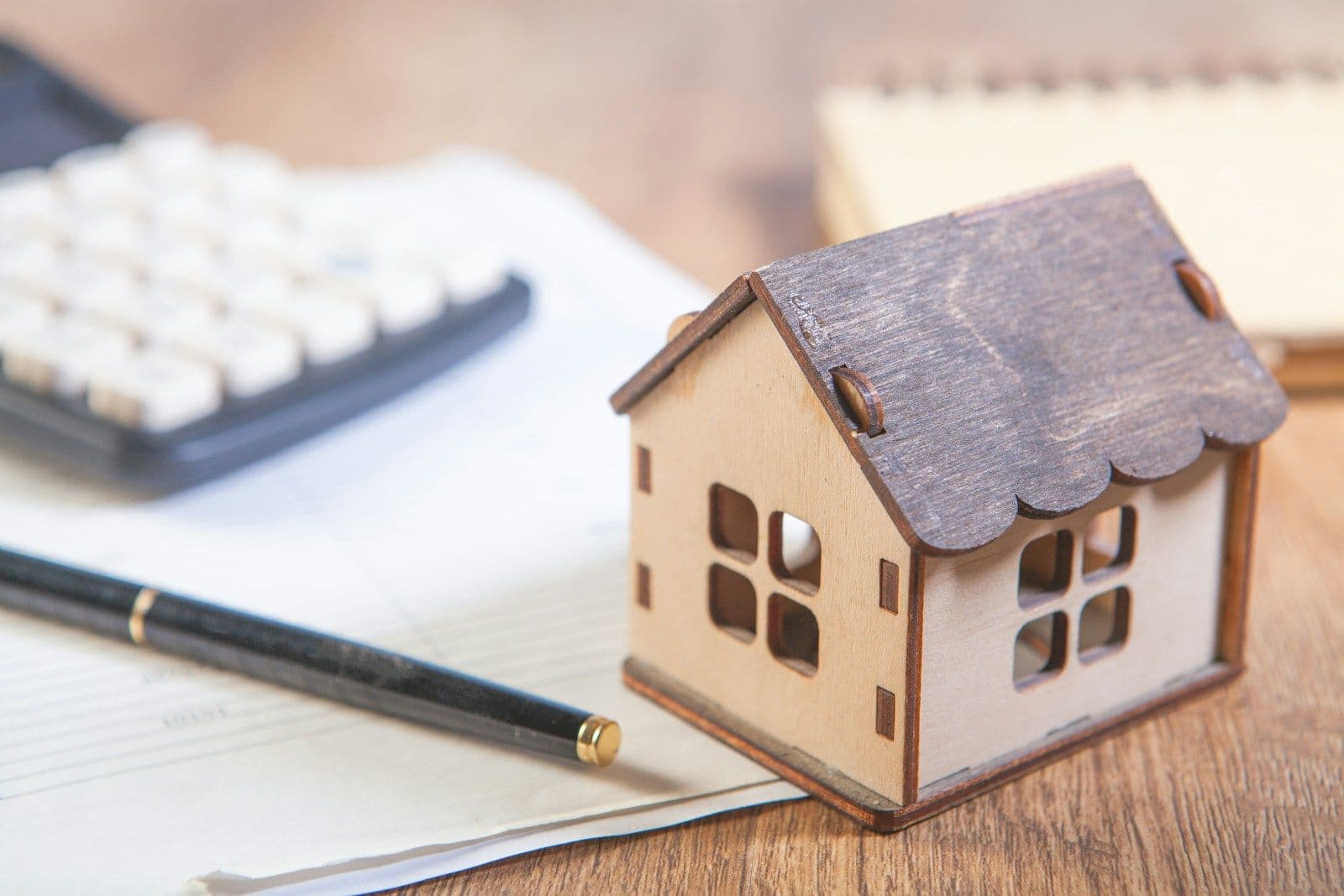 Small wooden toy house model beside a calculator, pen, and papers on a wooden desk, representing home finance