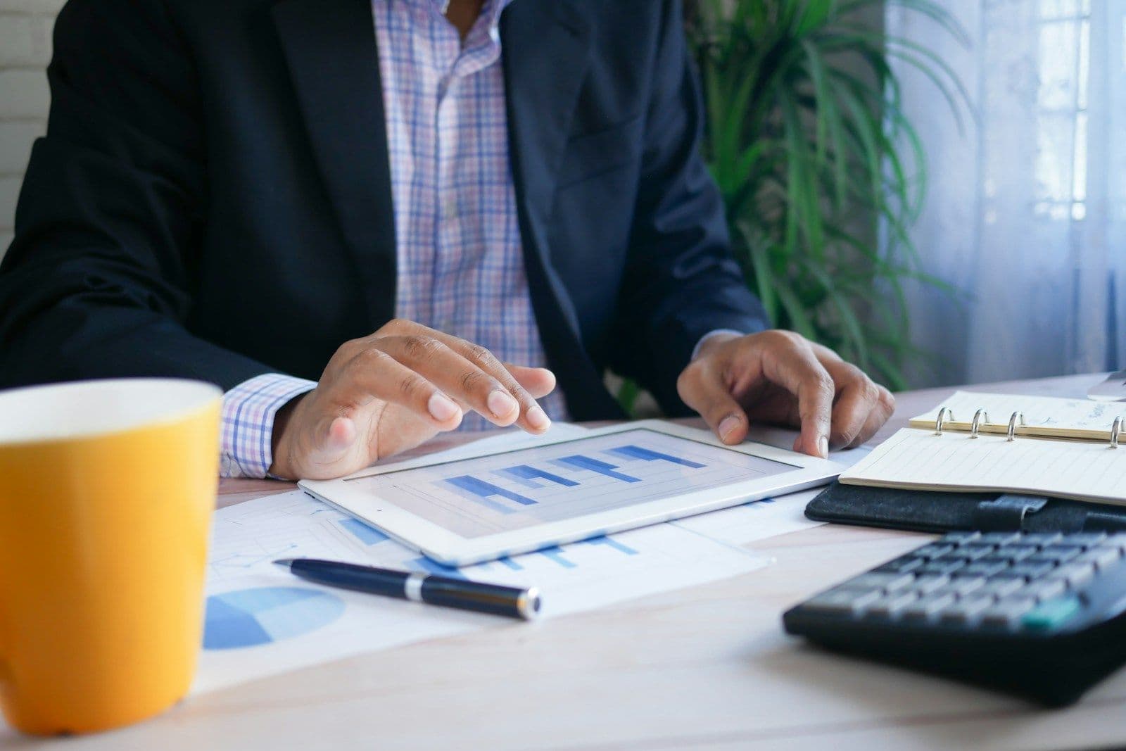 Person in a dark blazer tapping a tablet showing bar charts on a desk beside a calculator, pen, notebook, and yellow mug
