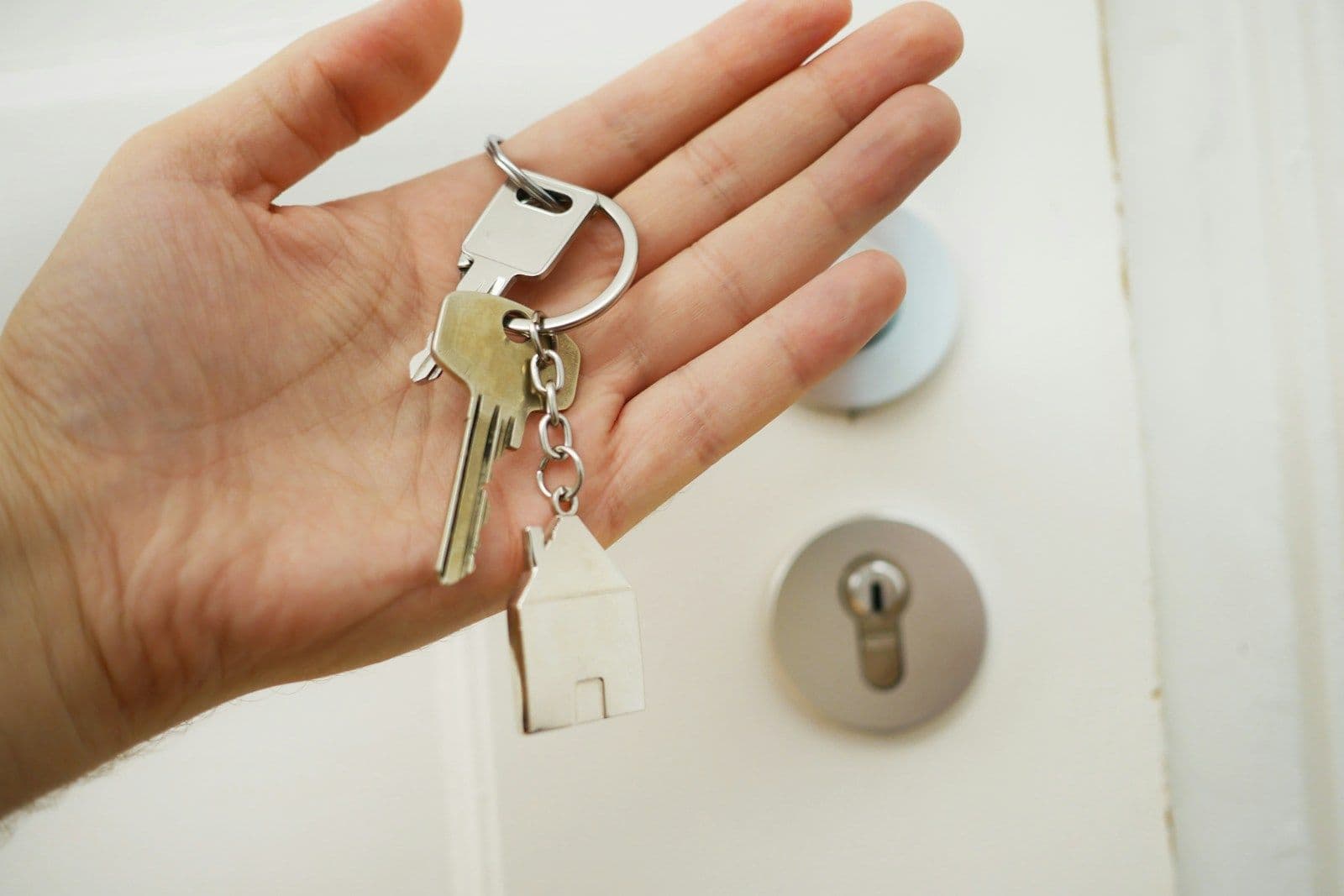 A hand holding a set of silver keys on a ring with a small white house-shaped keychain, with a white door and deadbolt lock in the background.