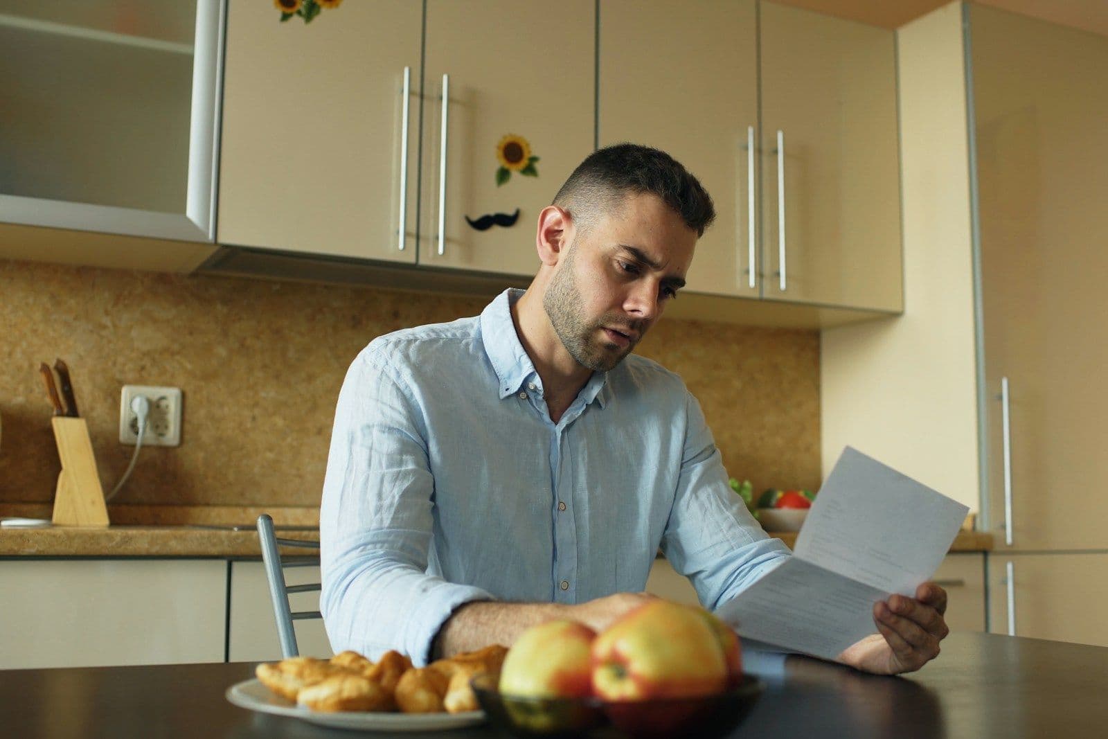 A man in a light blue shirt sits at a kitchen table reading a letter with a serious, concentrated expression.