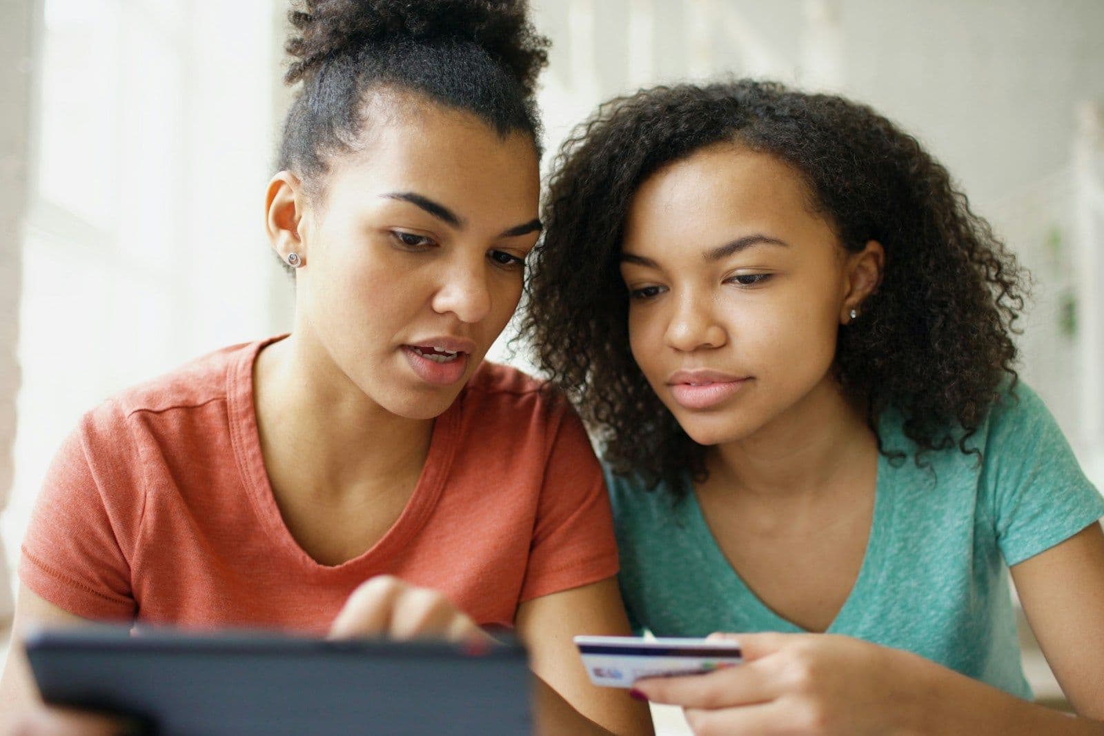 Two young women sit side by side looking at a tablet while one holds a credit card, appearing to make an online purchase or payment.