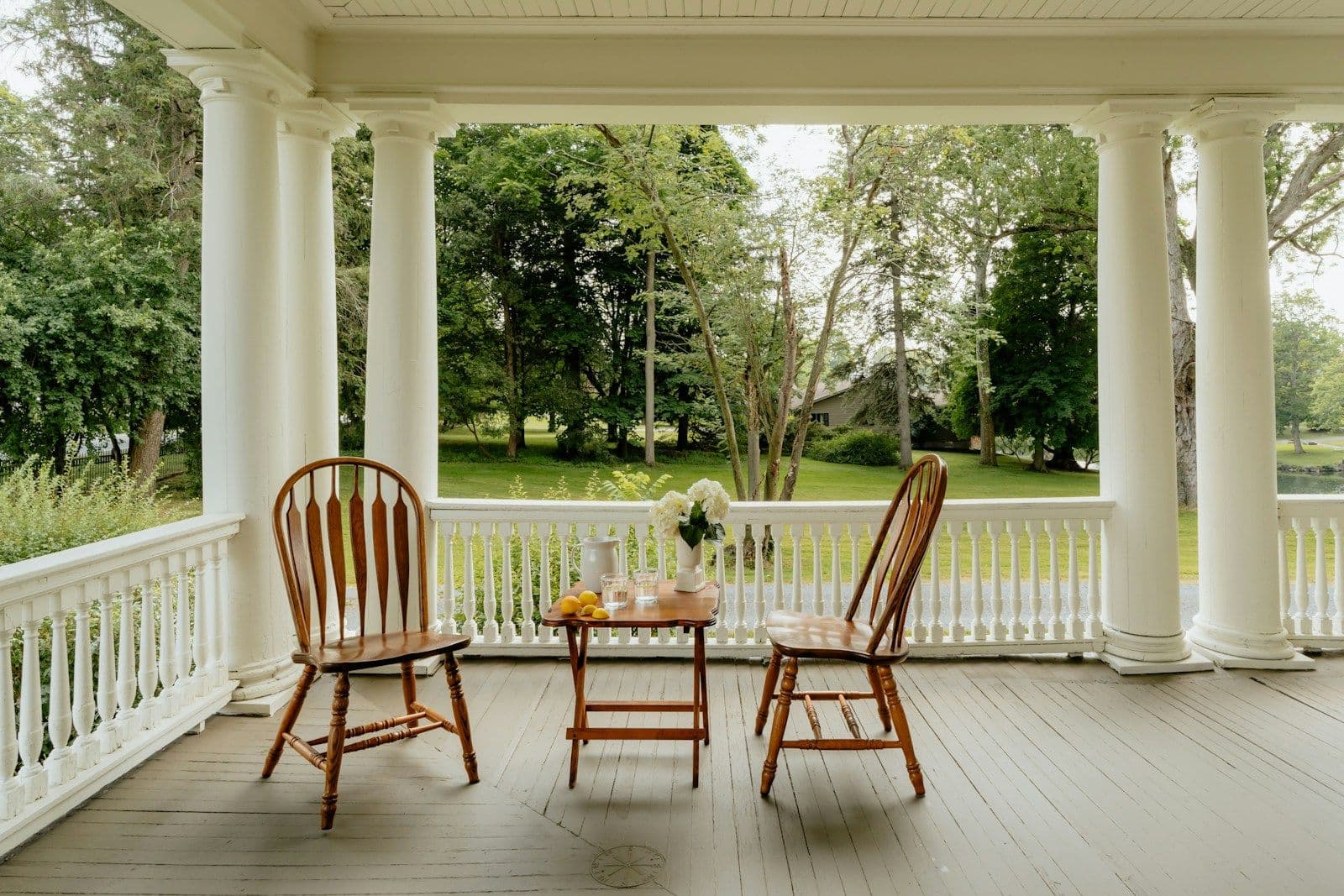 Two wooden chairs and a small table on a quiet white-columned porch overlooking green trees.