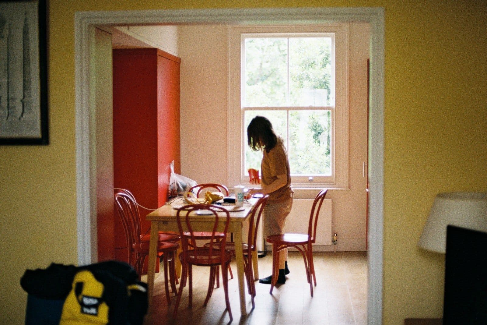 A woman stands at a wooden dining table by a bright window in a quiet, warmly lit home interior.