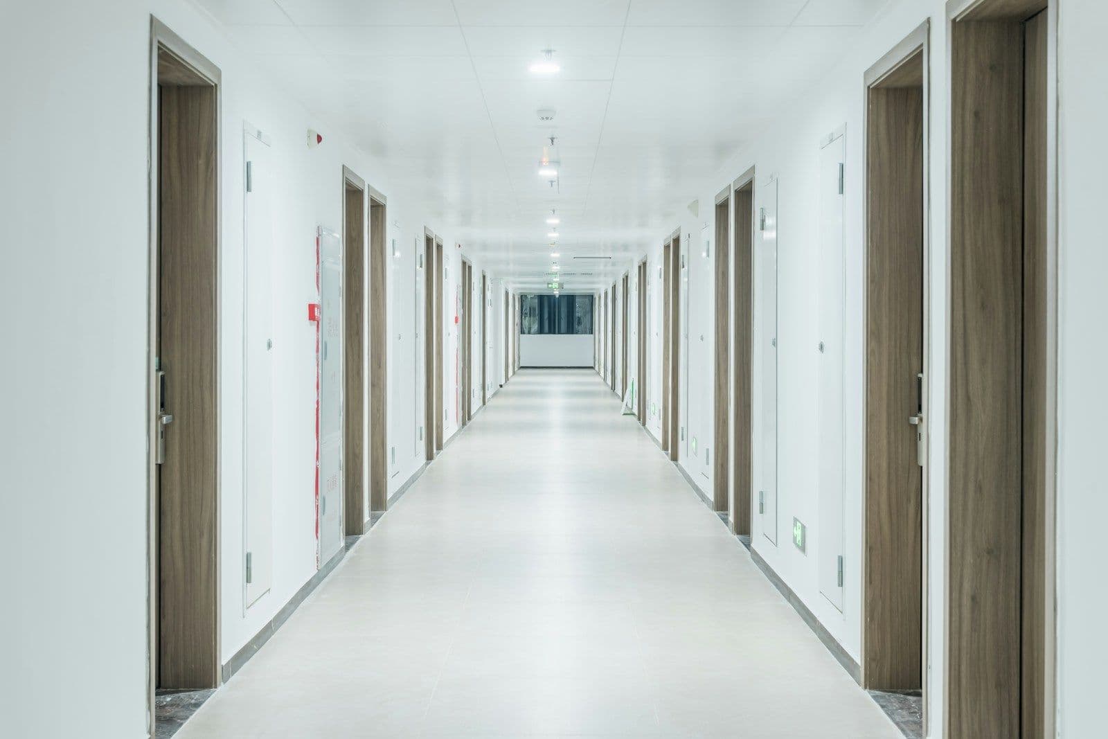 A long, bright, empty corridor with white walls and wooden doors leading toward a window at the far end.