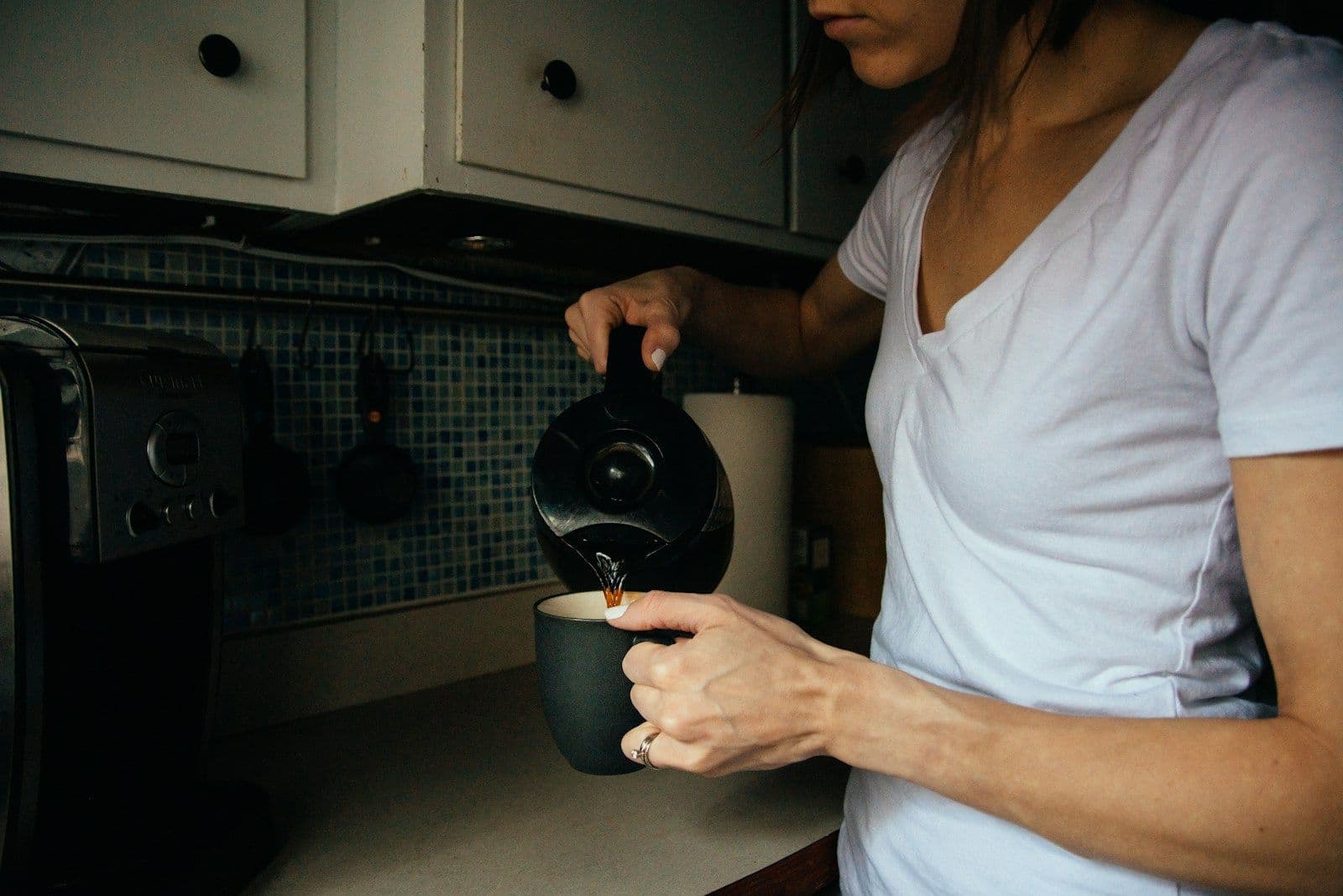A woman in a white t-shirt pours coffee from a carafe into a dark mug in a quiet kitchen.