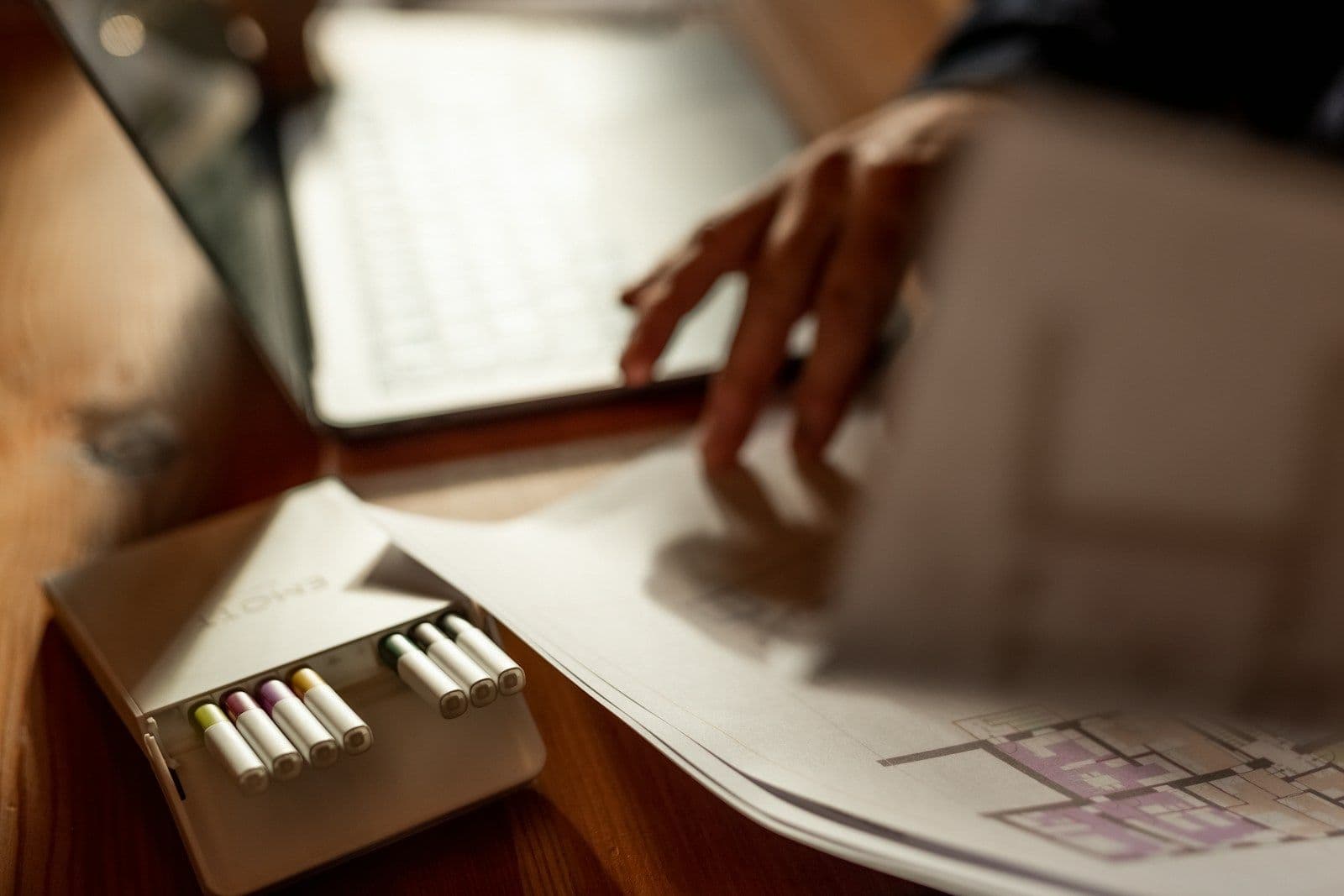 A person’s hands typing on a laptop beside an open pack of cigarettes and printed paperwork on a wooden desk