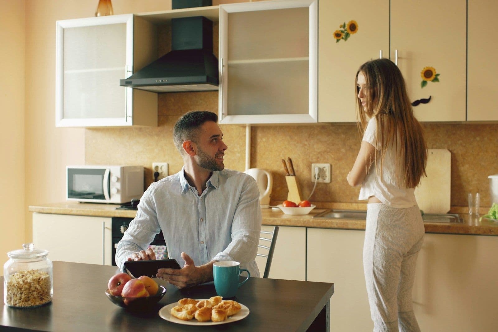 A man sits at a kitchen table looking at a tablet while a woman stands nearby in a bright, tidy home kitchen.