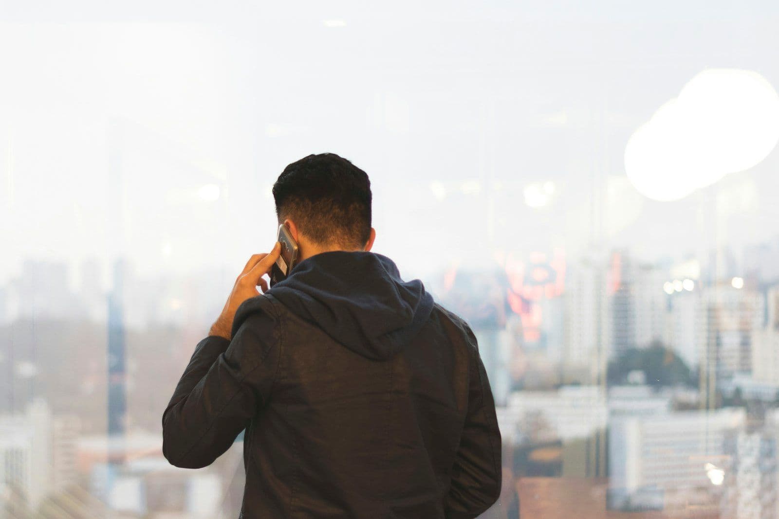 Man in a dark hooded jacket seen from behind, holding a phone to his ear while looking out a large window over a hazy city skyline.