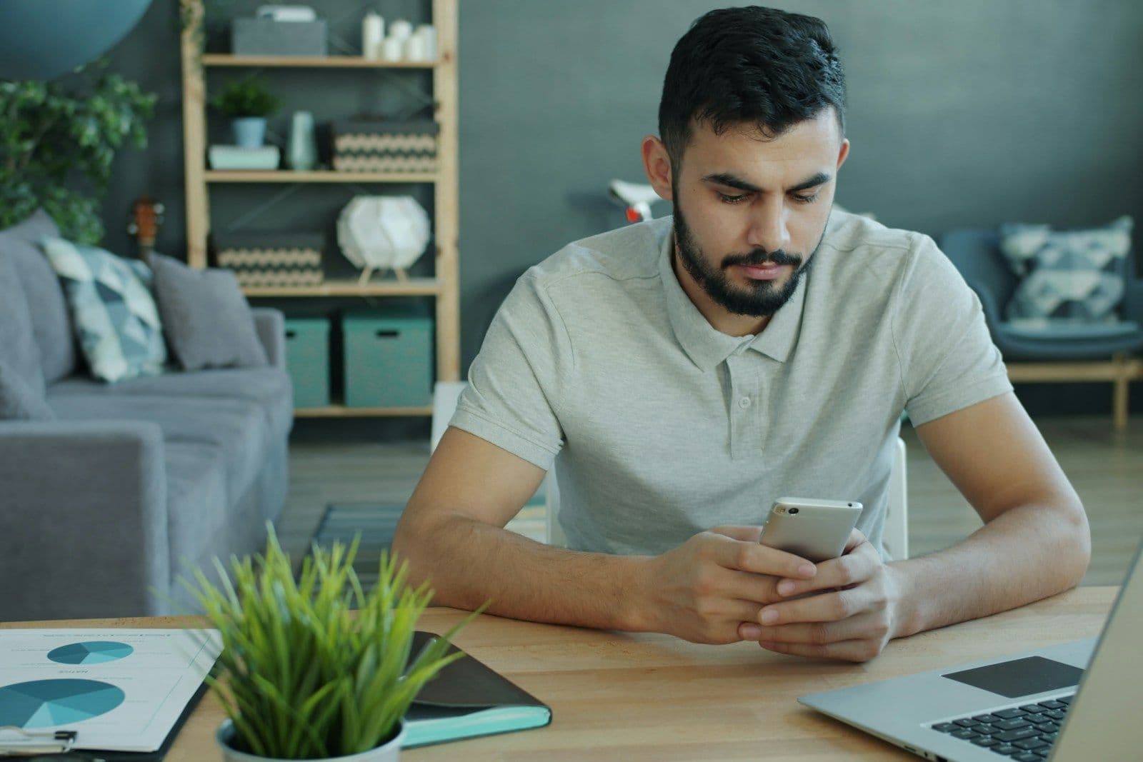 A young man in a gray polo shirt looks down intently at his smartphone while seated at a wooden desk beside an open laptop, a potted plant, and a chart.