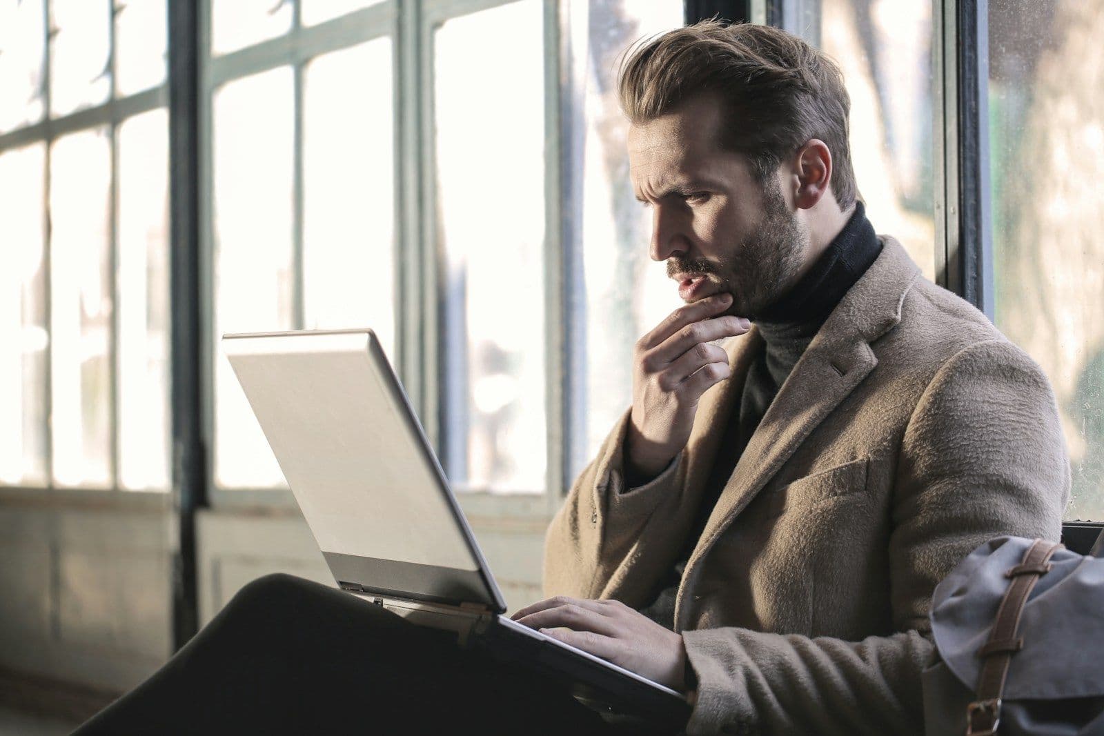 A bearded man in a tan coat and dark turtleneck sits by a window, looking intently at an open laptop while resting his hand on his chin.