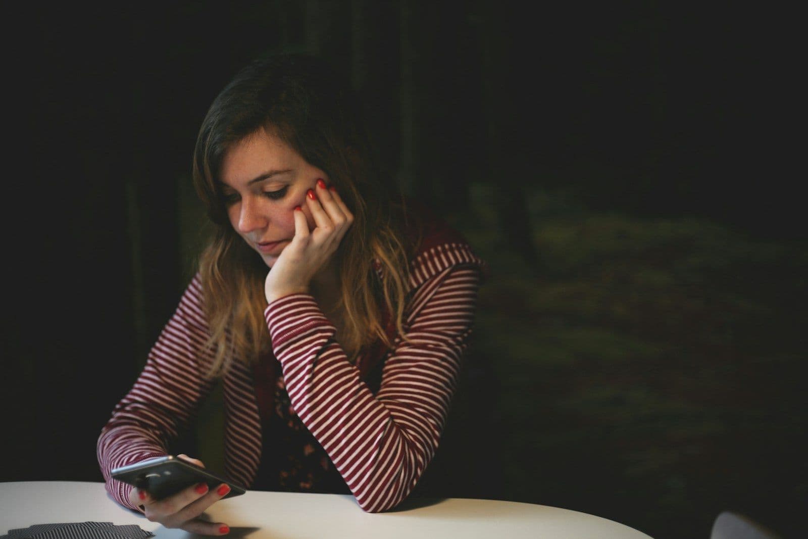 A woman in a red and white striped top sits at a table in a dark room, resting her chin on her hand while looking down at a smartphone.