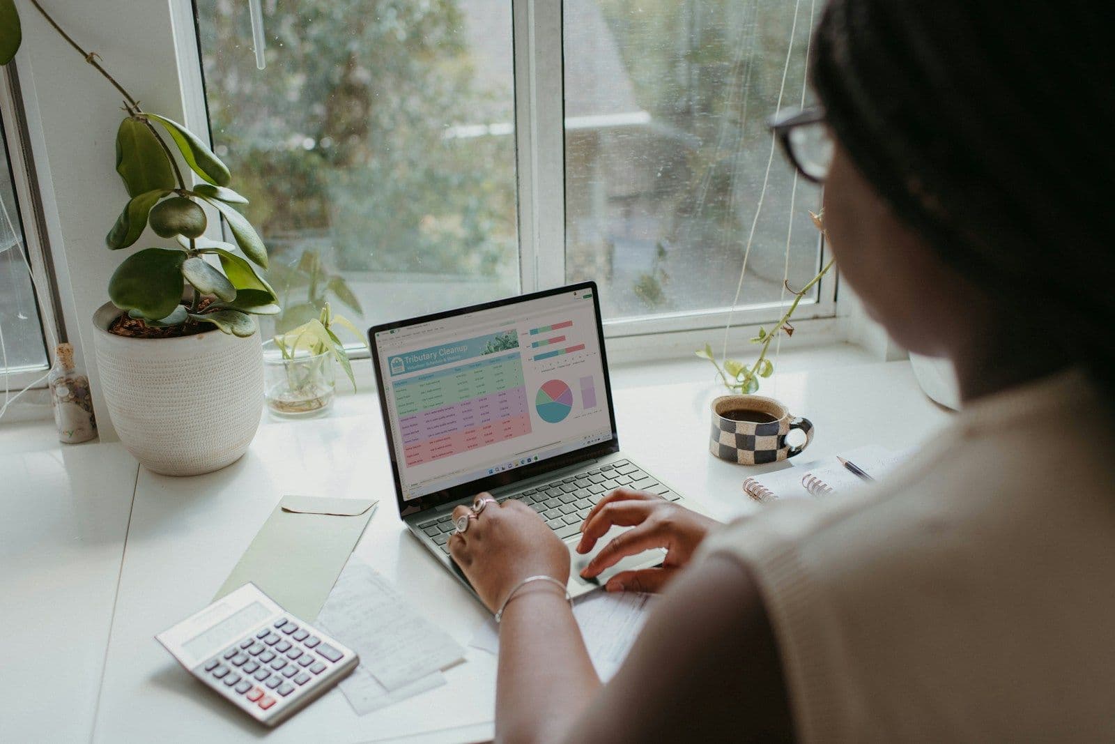 Person with glasses working on a laptop at a white desk by a window, with a calculator, notepad, and coffee mug nearby