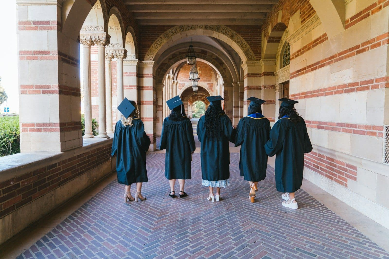 Five graduates in caps and gowns walk away from the camera down a brick arcade with arched columns.