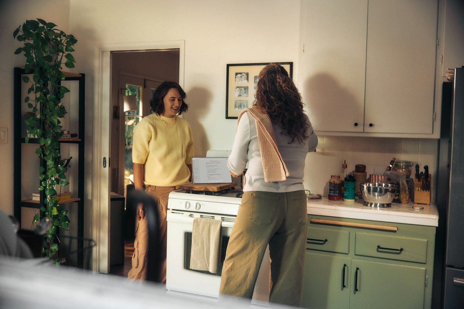 Two housemates chat and smile in a bright shared kitchen with green cabinets.