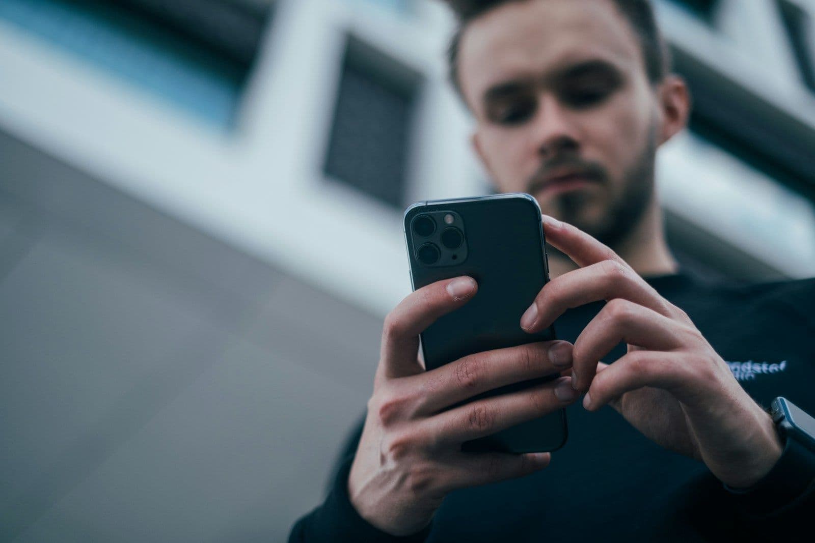 A young man with short hair and stubble looks down intently while typing on a black smartphone, shot from a low angle against a blurred city building.