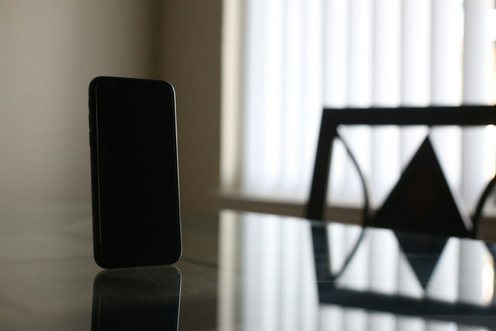 A dark smartphone standing upright on a glossy glass table, with a blurred triangular ornament and window blinds in the background