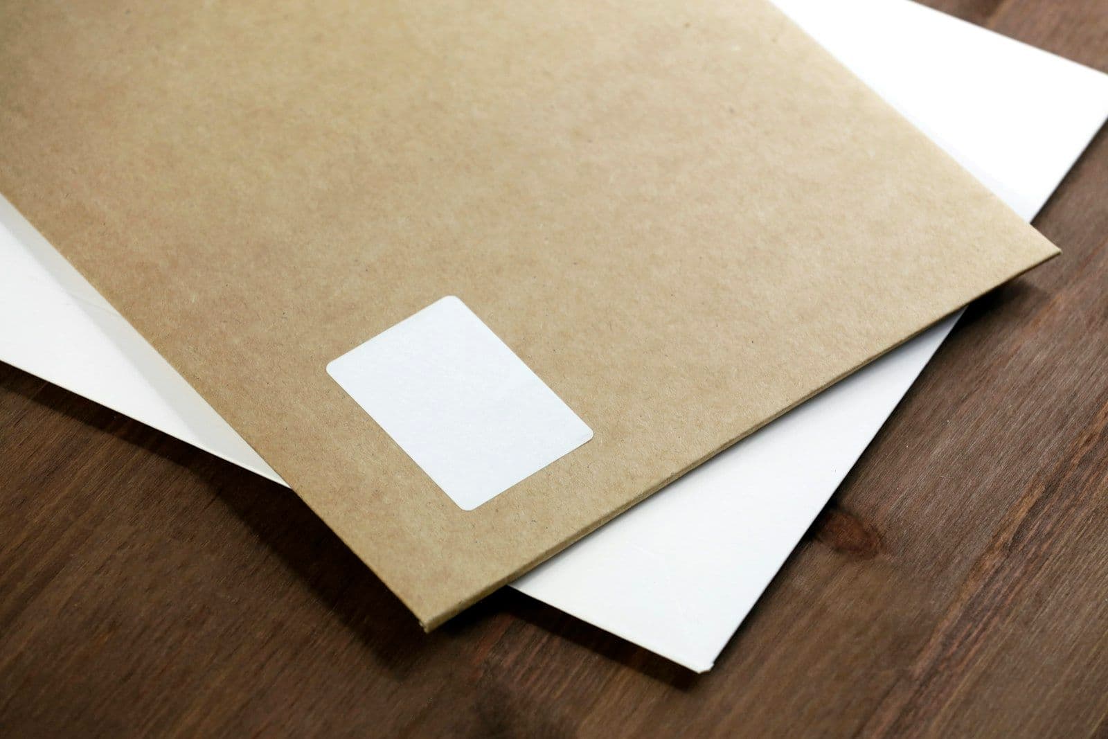 A brown kraft paper envelope with a blank white address label rests on top of a white sheet of paper on a dark wooden table.