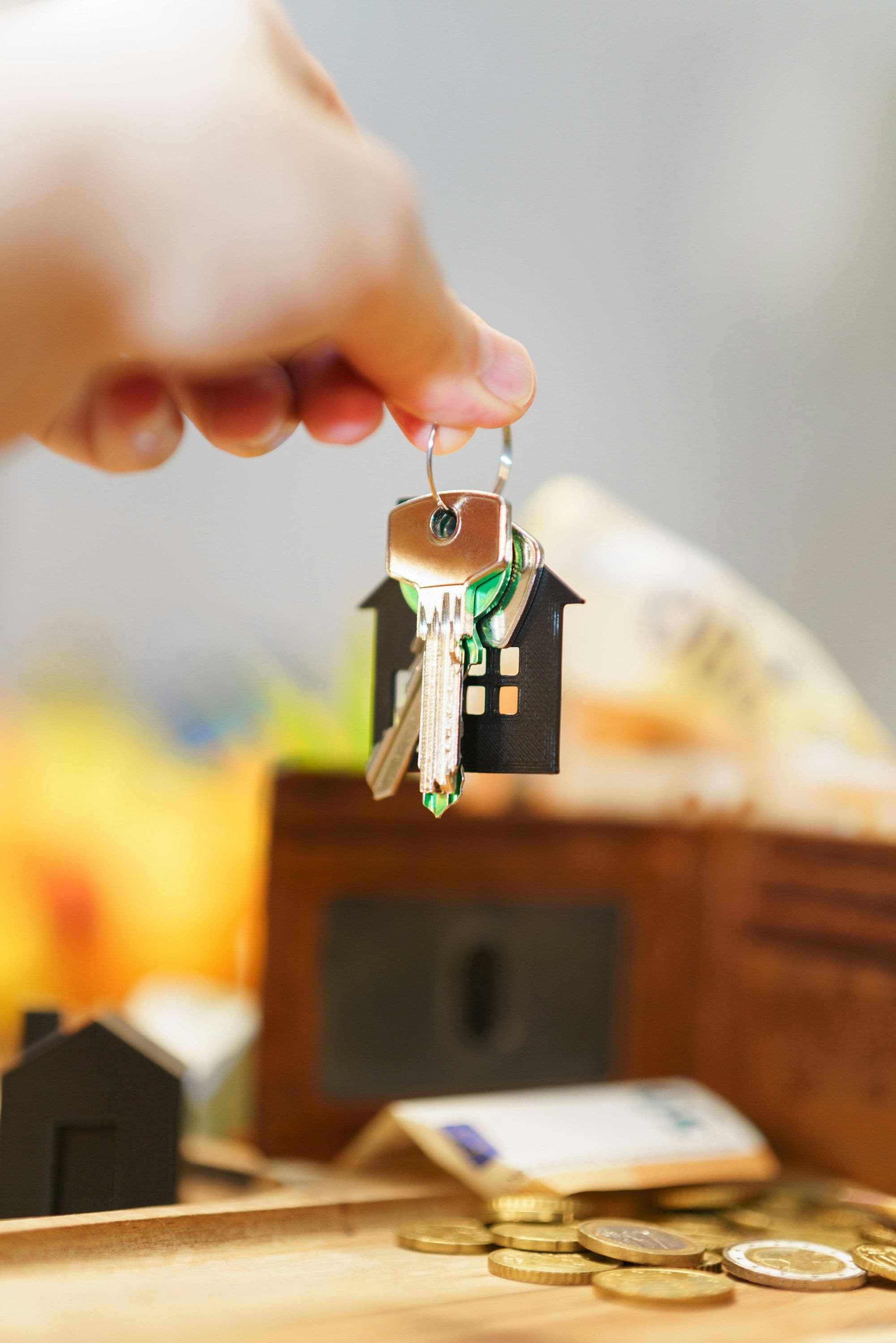 Hand holding keys with a small house-shaped keychain above coins and a wooden surface.