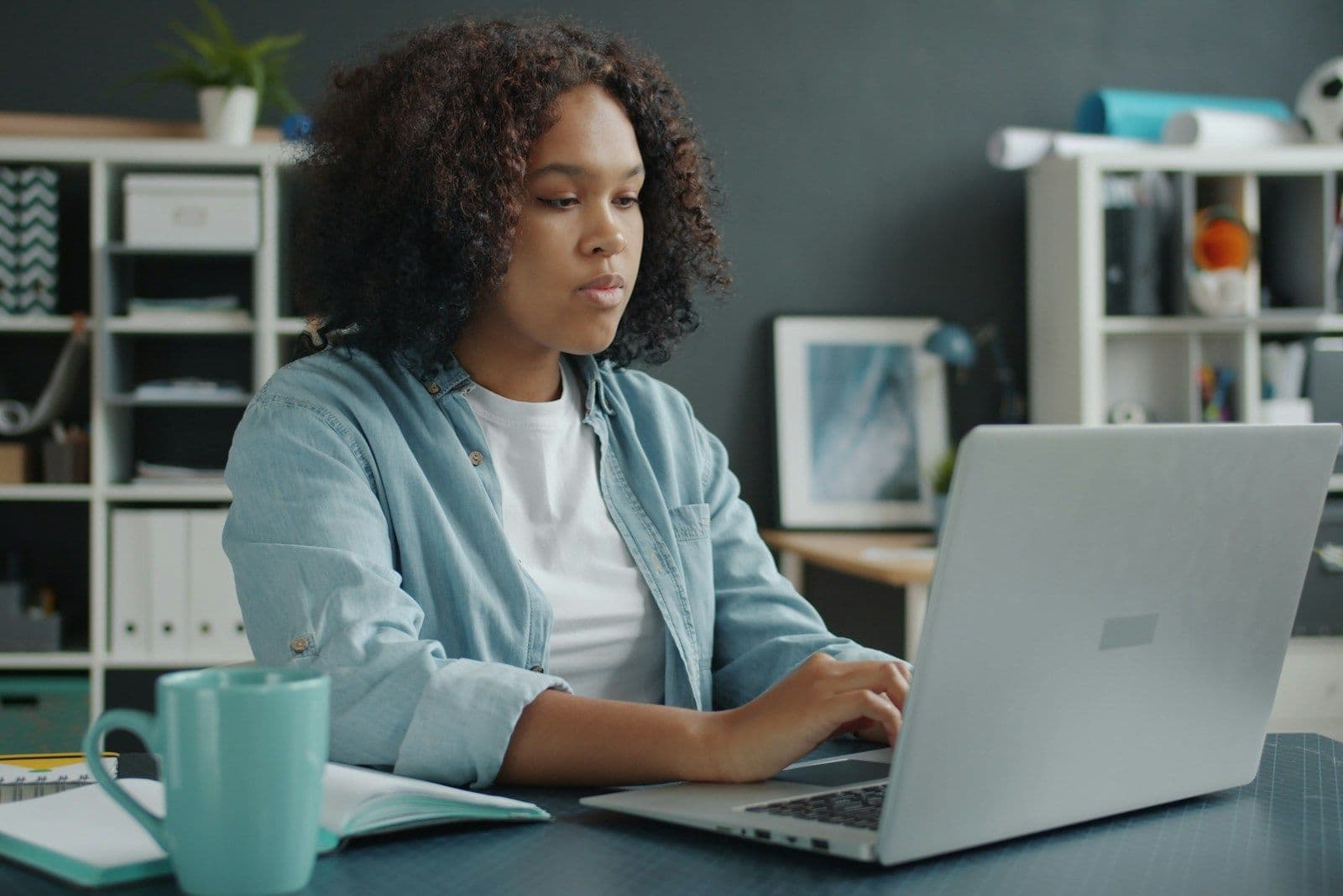A young woman with curly hair in a light blue shirt types on a silver laptop at a desk with a teal mug, notebook, and shelves in a home office.