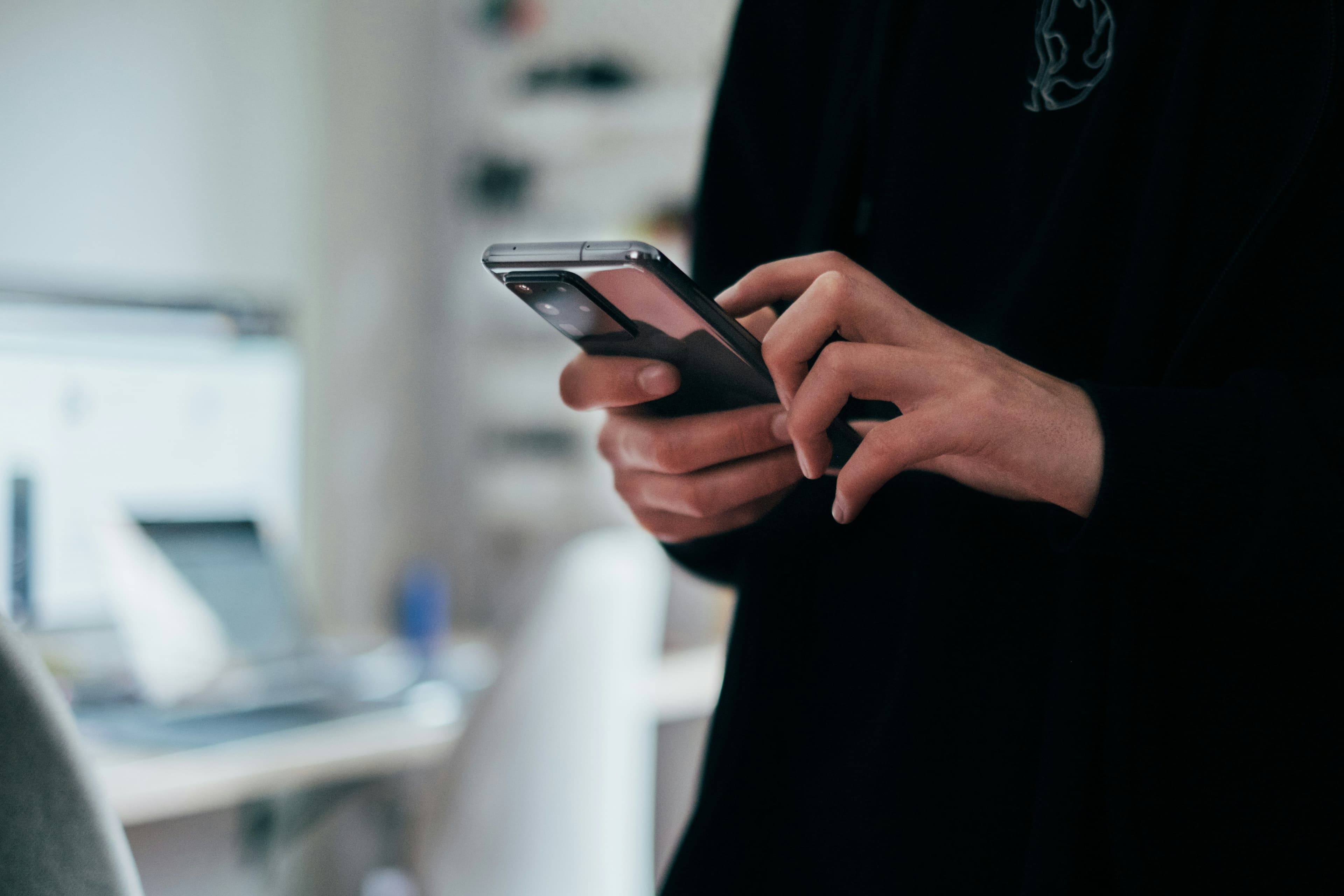 Close-up of hands holding and tapping a smartphone, with a blurred laptop in the background.