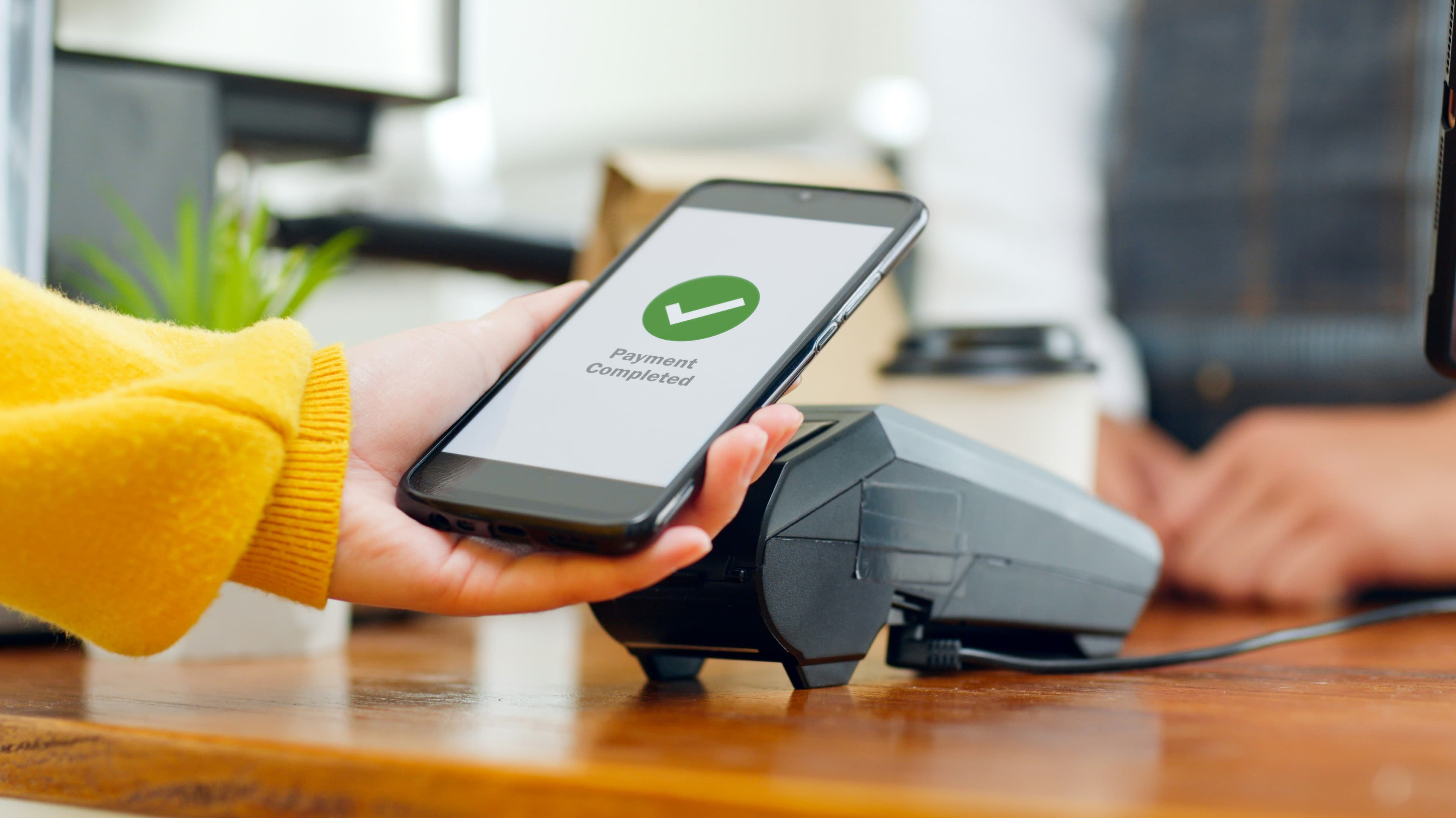 Hand in yellow sweater holding a phone showing "Payment Completed" above a card terminal on a wooden counter.