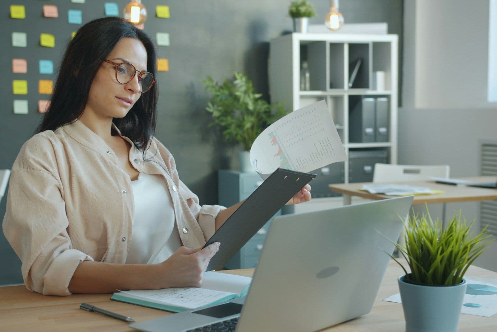Woman with glasses reviewing papers in a black folder at a desk with an open laptop, notebook, and potted plant in a bright office