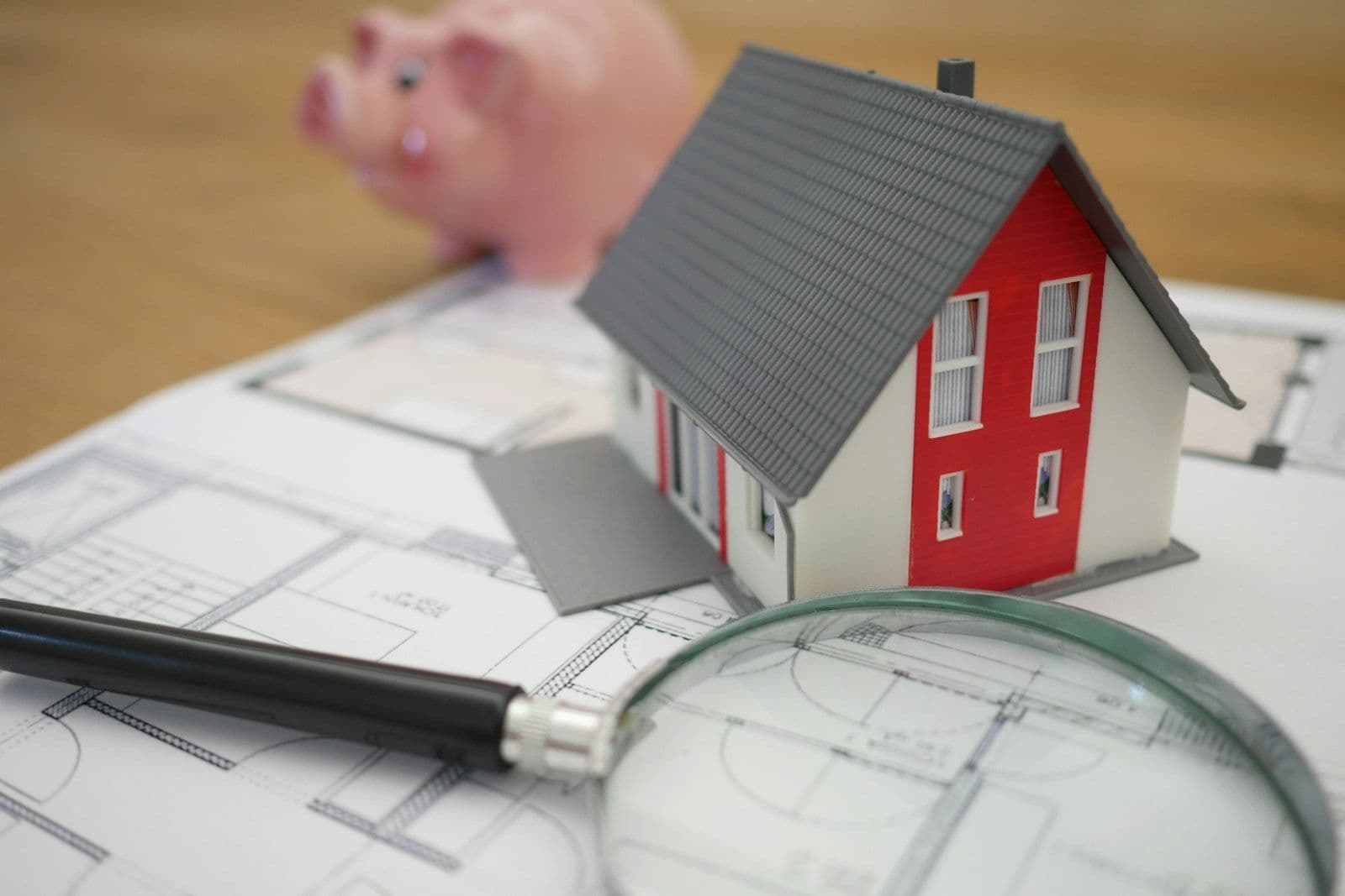 Miniature red house model with gray roof resting on blueprint floor plans, with a magnifying glass and a pink piggy bank in the background
