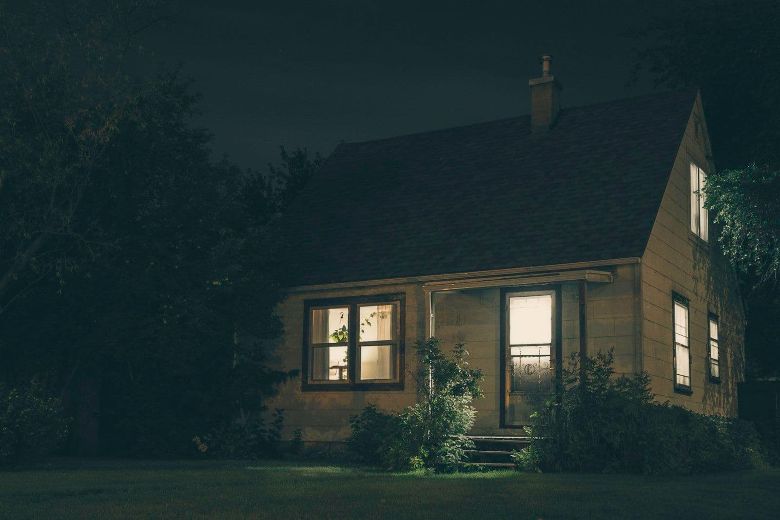 A small wood-sided cottage at night with warm light glowing from its windows, surrounded by dark trees and shrubs on a grassy lawn.