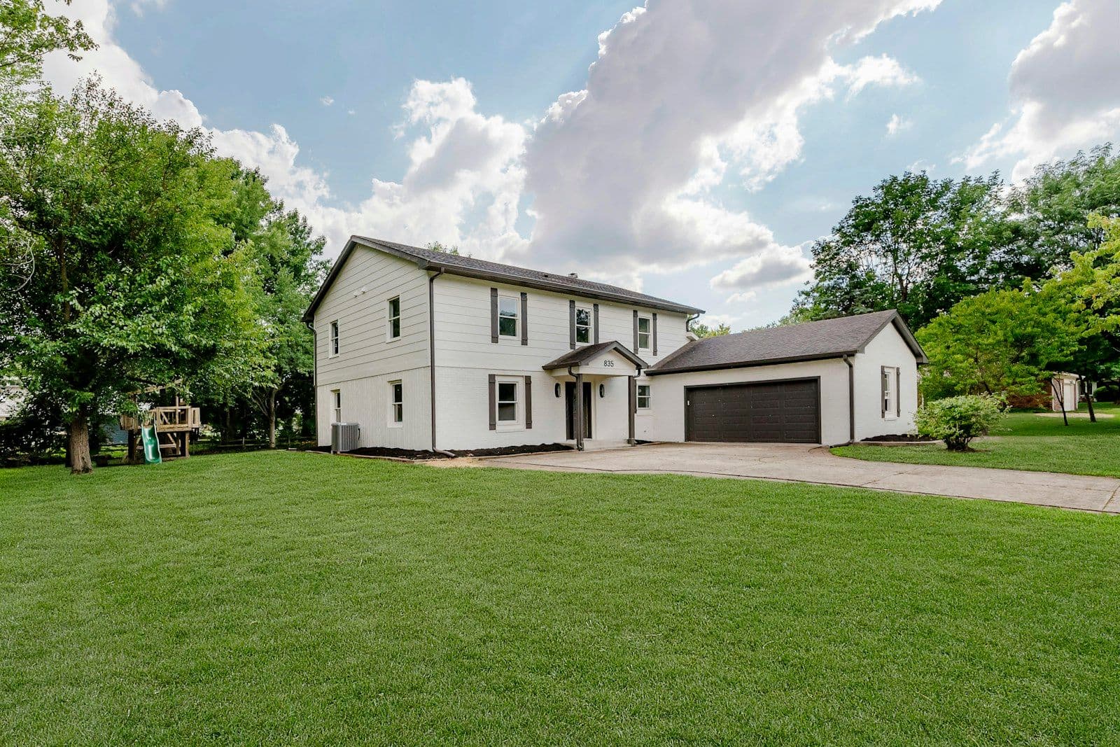 Two-story white house with black shutters, a covered front porch, and an attached dark garage, set on a green lawn under a partly cloudy sky.