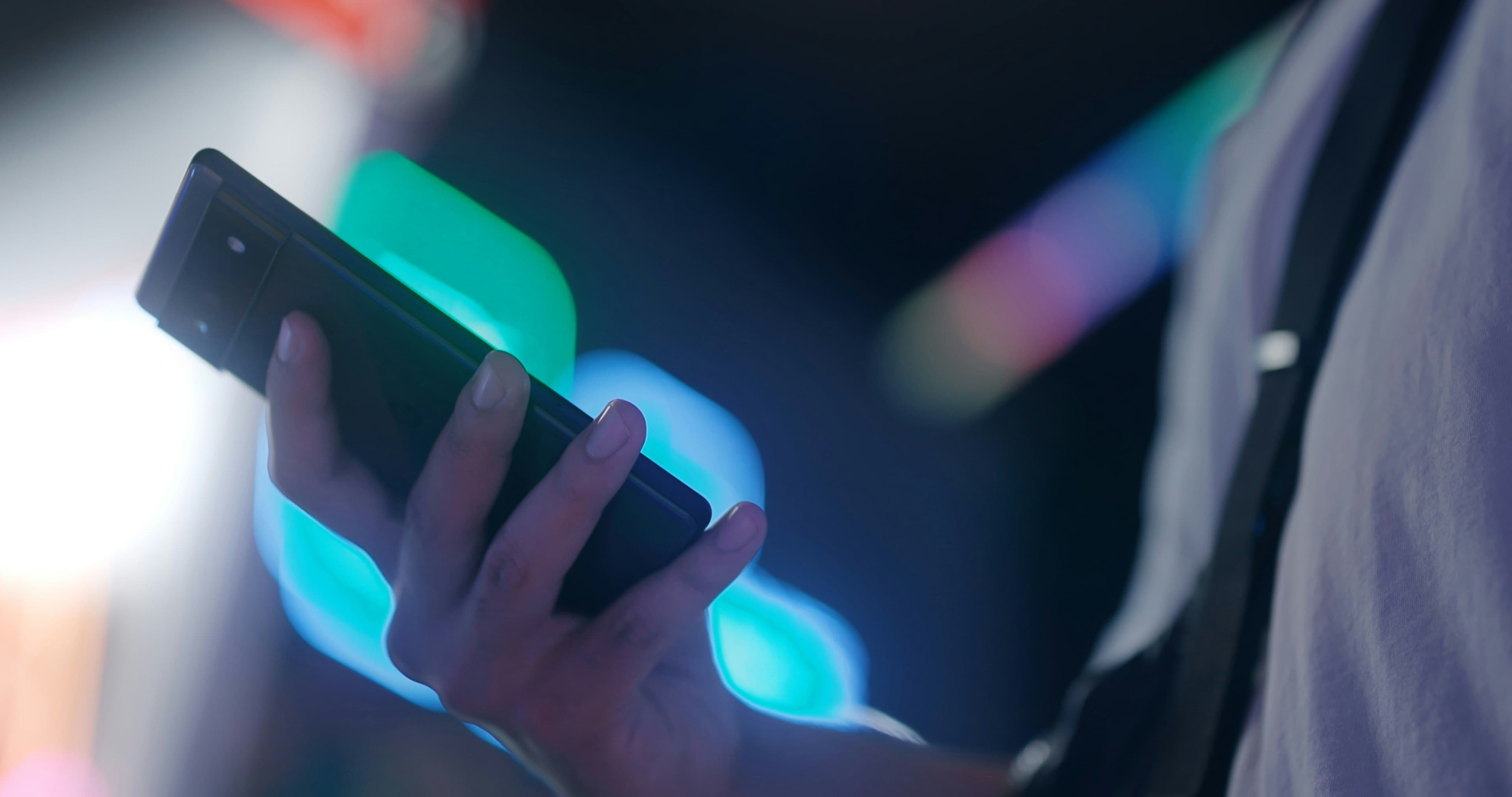 Hand holding a smartphone against a dark background with green and blue neon light bokeh.