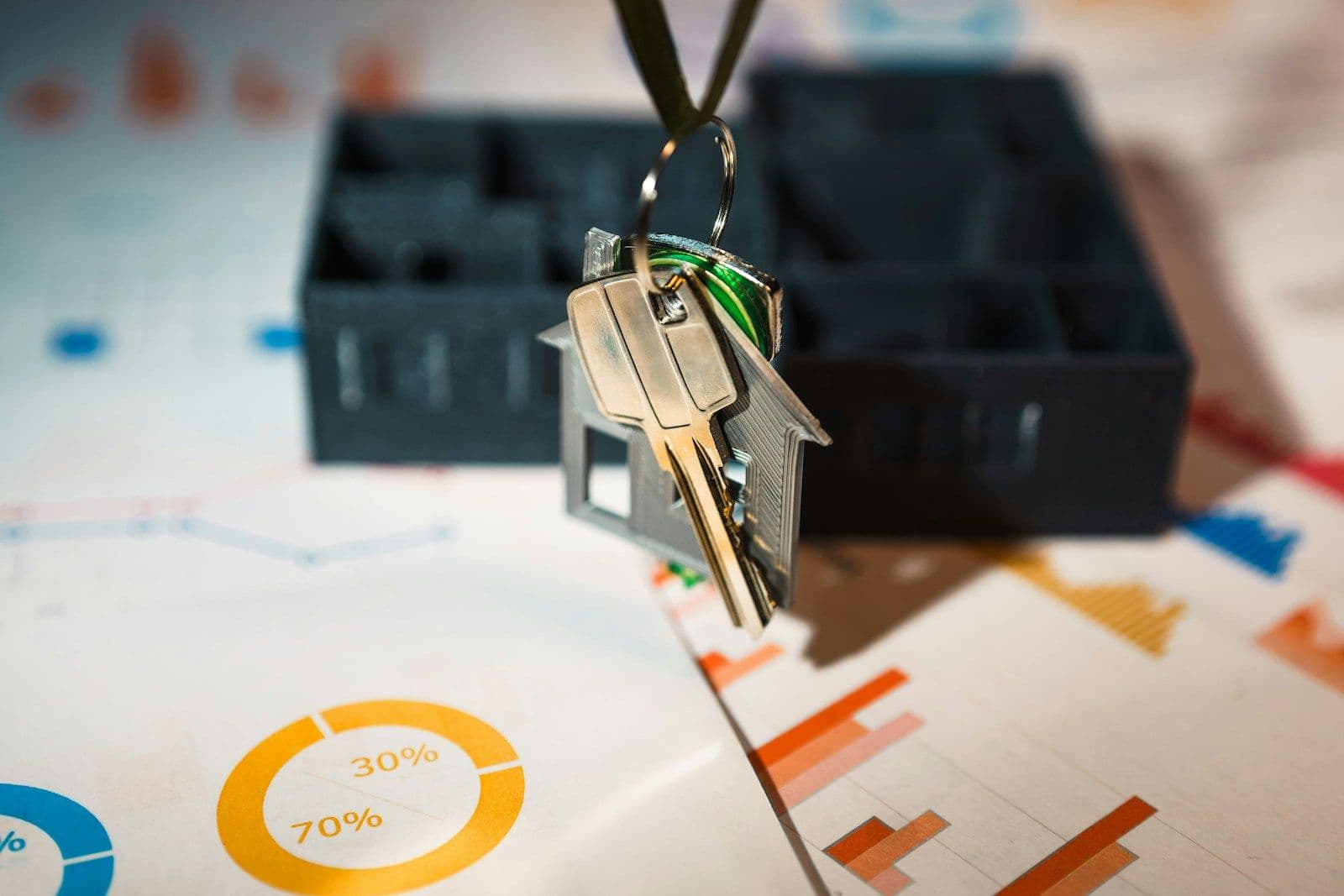 A set of house keys with a small metal tag hangs in front of a black organizer tray, over financial charts with pie graphs and bar graphs.
