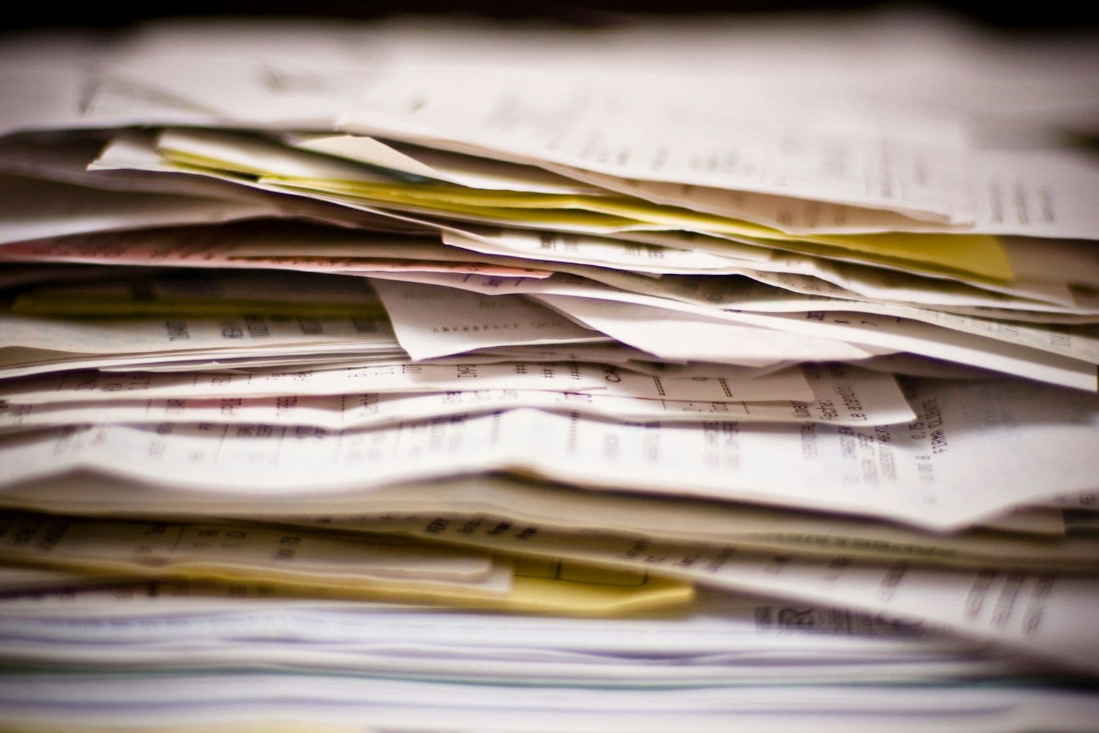 A tall, blurred stack of paper documents, letters, and bills with yellow and white sheets fanned out, suggesting piles of debt-related correspondence.