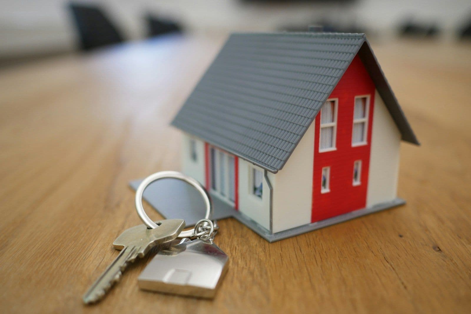 Small model house with a gray roof and red door on a wooden table, with a set of keys and a house-shaped keychain in front