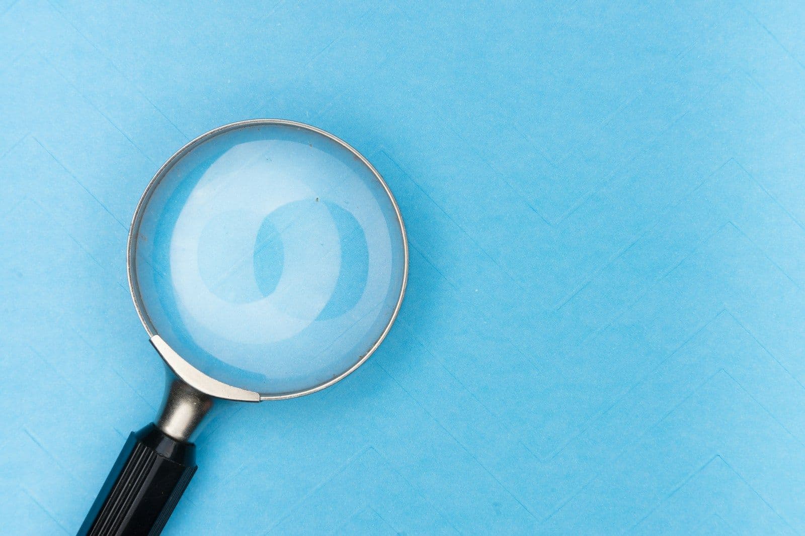 A magnifying glass with a black handle lying on a light blue background, with a faint circular reflection visible through the lens.
