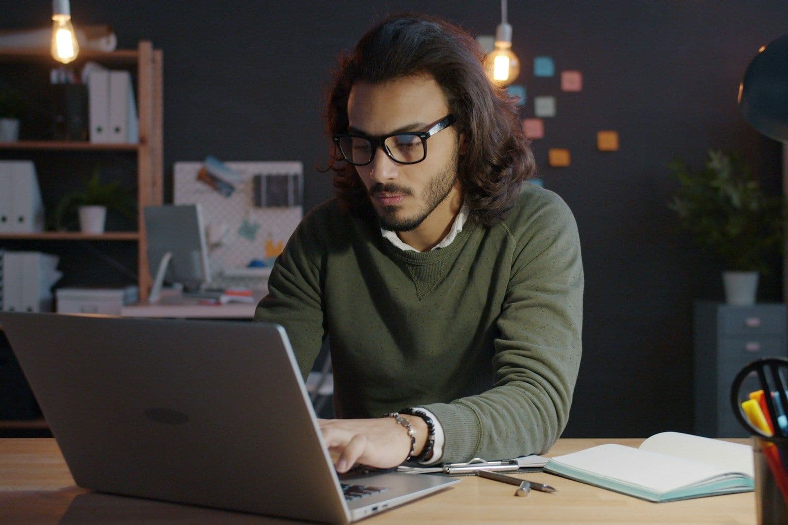 A man with glasses works on a laptop at a desk in a dimly lit home office, with a notebook and pens nearby.