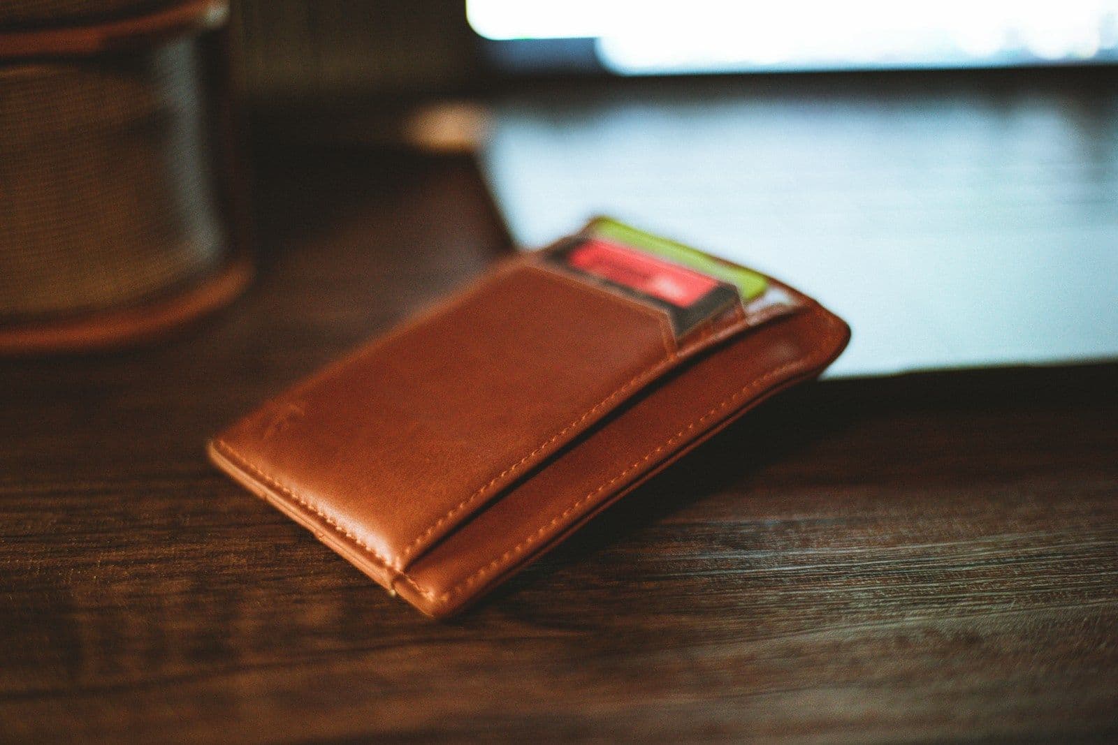A tan leather bifold card holder with stitched edges resting on a dark wooden table, with a few colorful cards tucked into its front pocket.