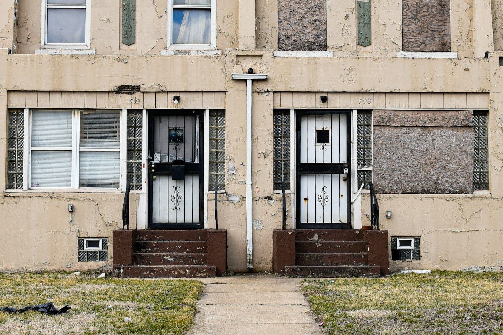 Two-story beige apartment building with two front doors, one boarded up with plywood, and dark brown entry steps and dead grass in front.