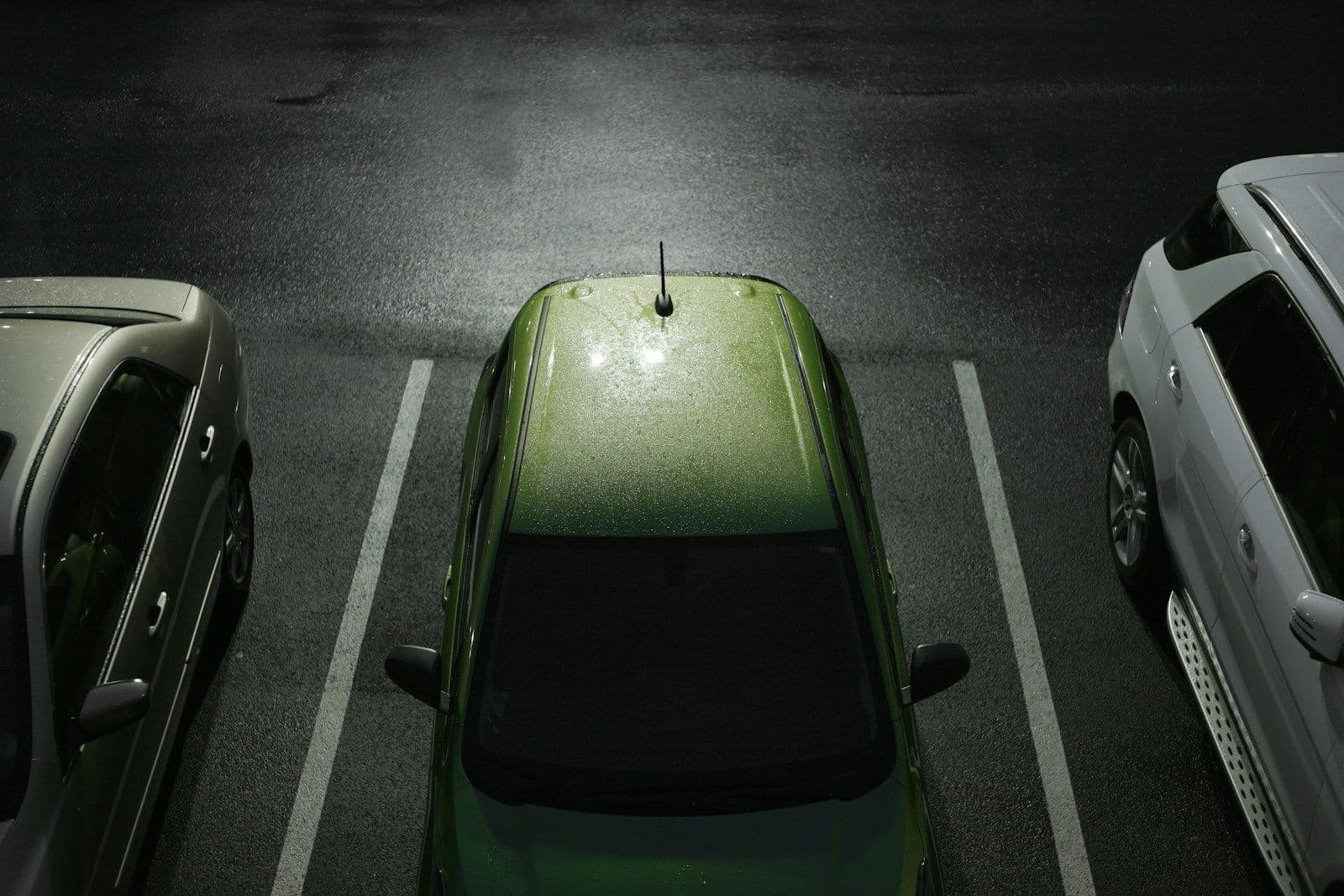 Overhead night view of a green hatchback parked between white lines in a wet parking lot, with silver cars on either side