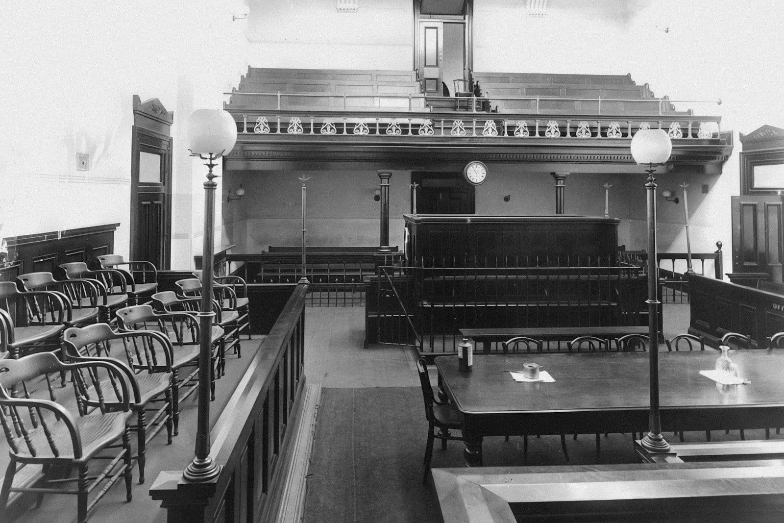 Black-and-white photo of an old courtroom interior with wooden spectator benches, a raised judge’s bench, and tables with lamps in the foreground.